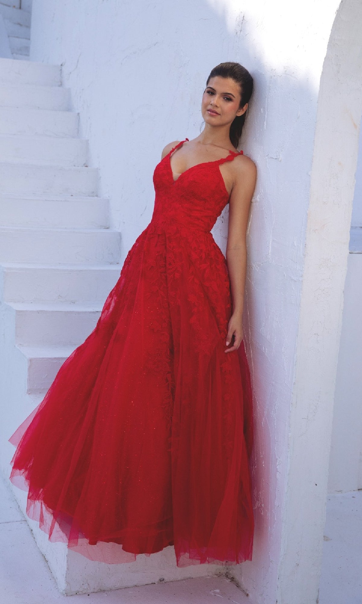A woman in the Embroidered-Lace Long Prom Dress: Eureka 9858, a bright red, floor-length v-neck gown with thin straps, stands by a white wall near white steps.