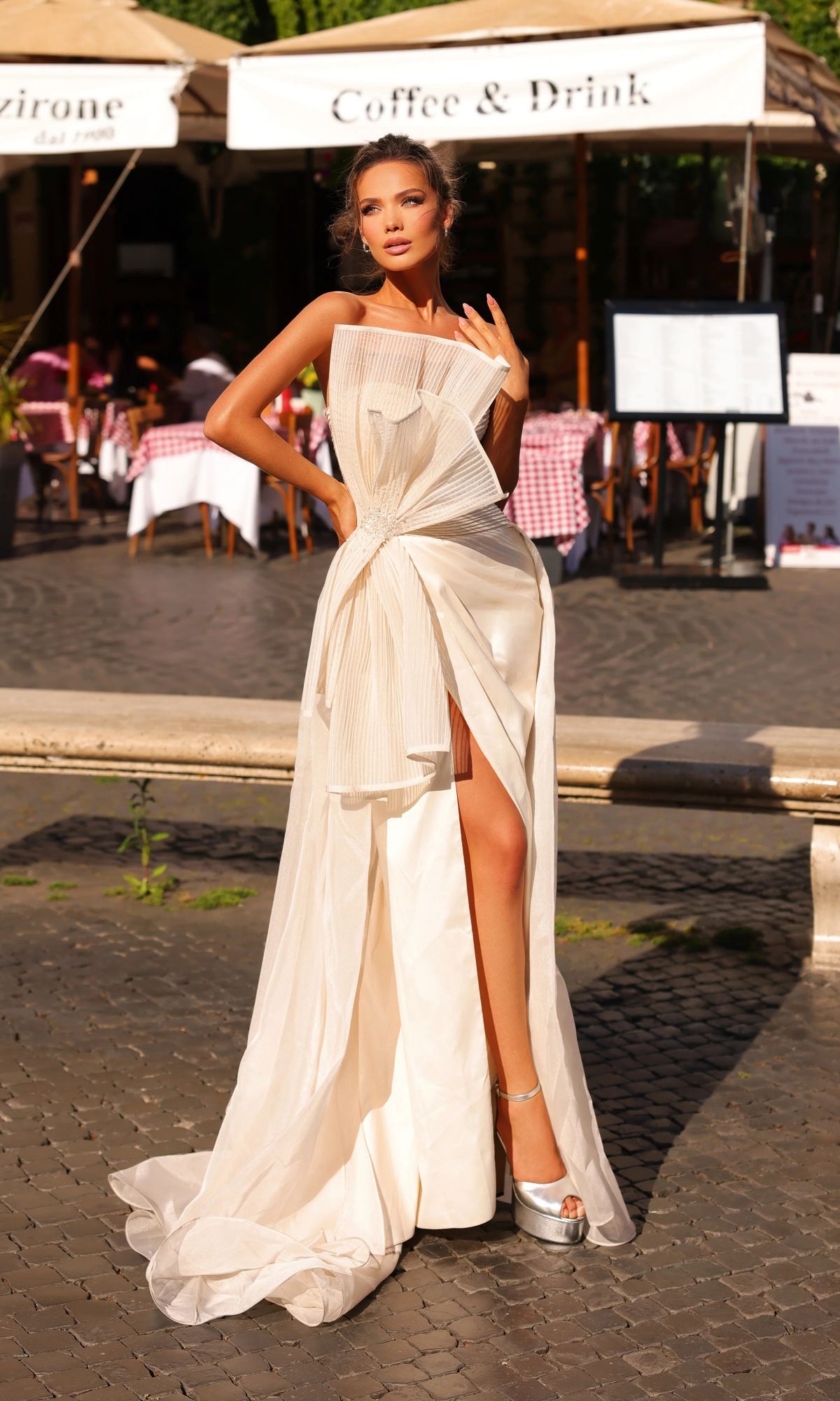 A woman in a vanilla Tarik Ediz 98708 long formal dress with a structured, strapless design and high slit poses on a cobblestone street near a café with outdoor seating.