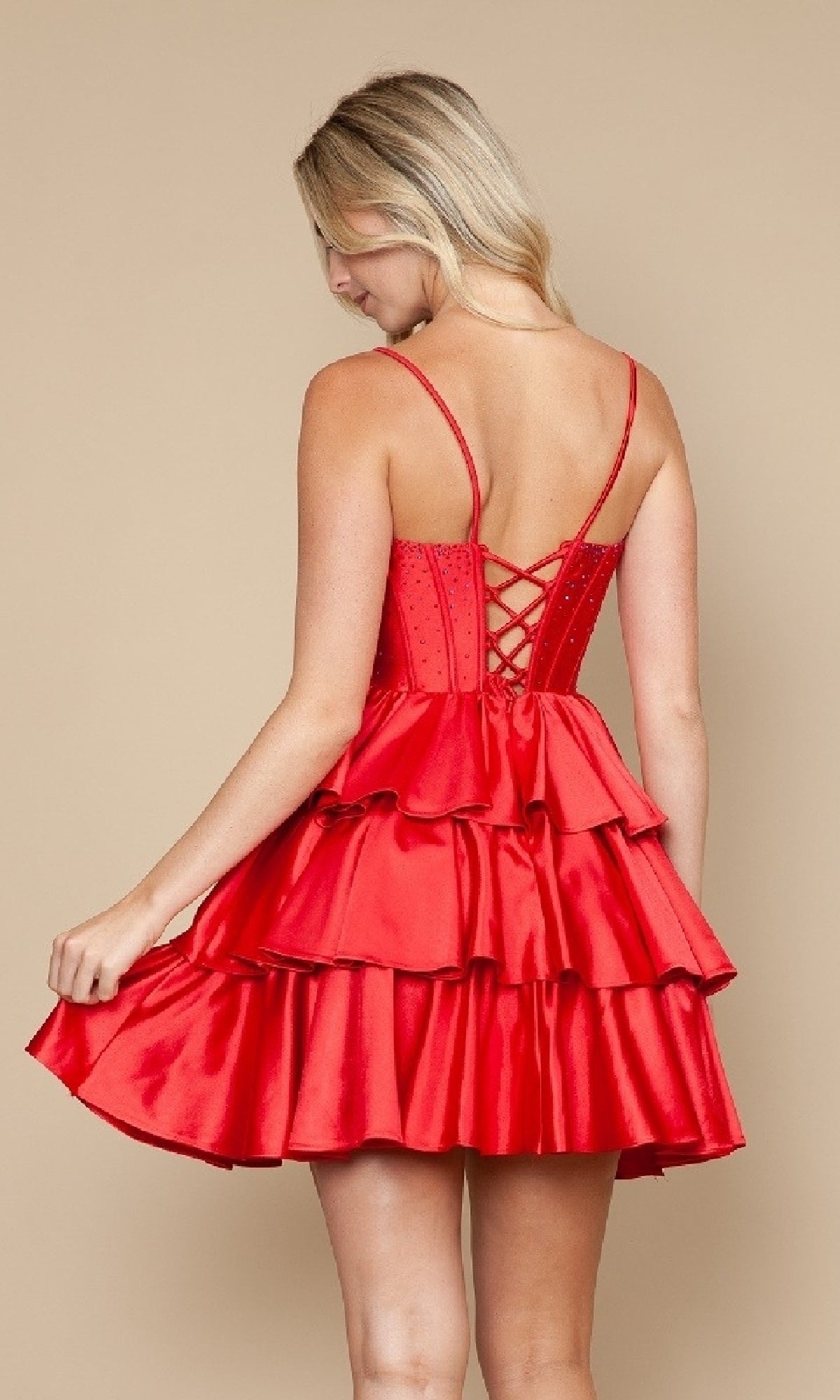 Woman modeling the Lace-Up Short Ruffle Hoco Dress: Poly USA 9896 in red with spaghetti straps, lace-up back, and tiered skirt, stands against a beige background facing away.