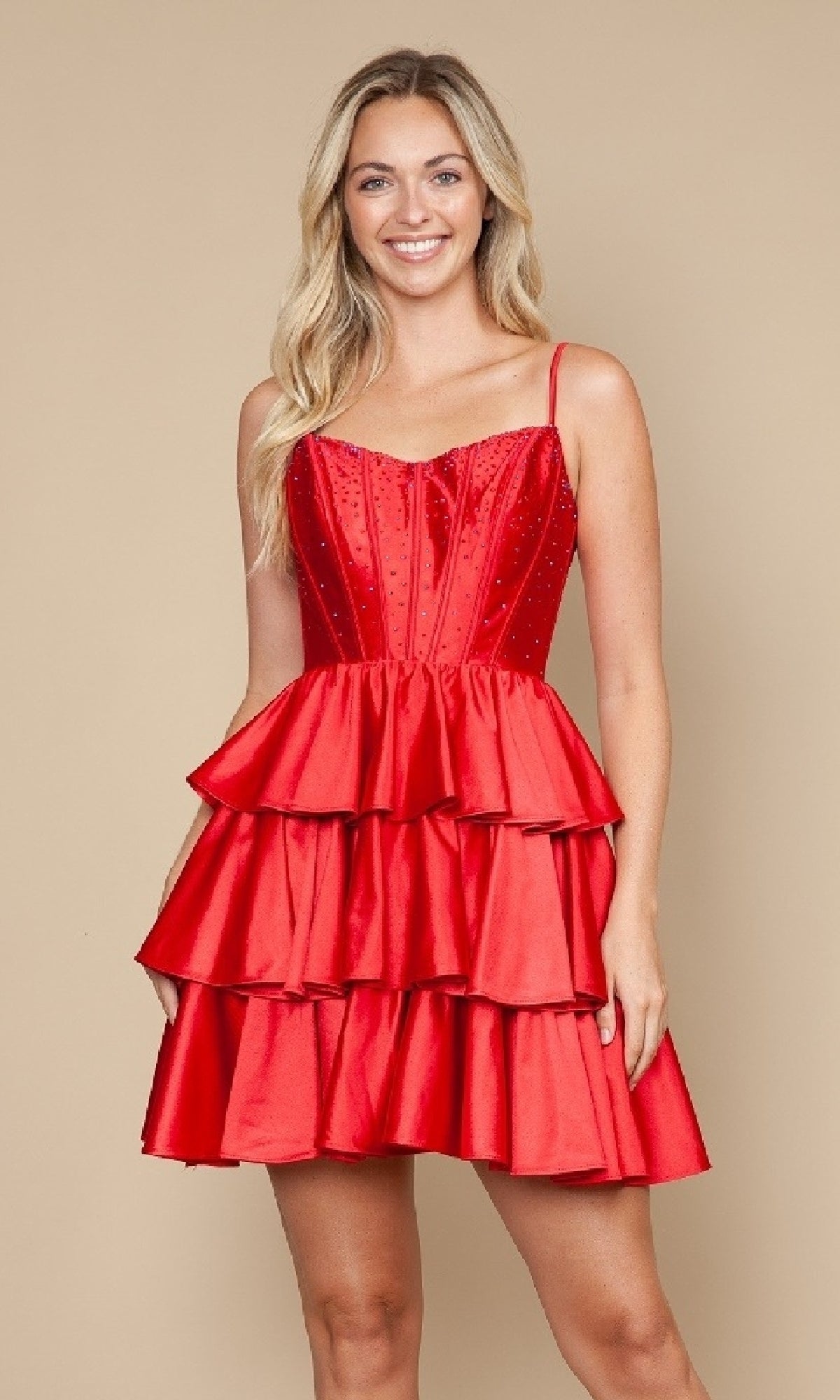A woman smiles while wearing the Poly USA 9896 Lace-Up Short Ruffle Hoco Dress, a sleeveless fit-and-flare red cocktail dress with thin straps and tiered ruffles, standing against a plain beige background.