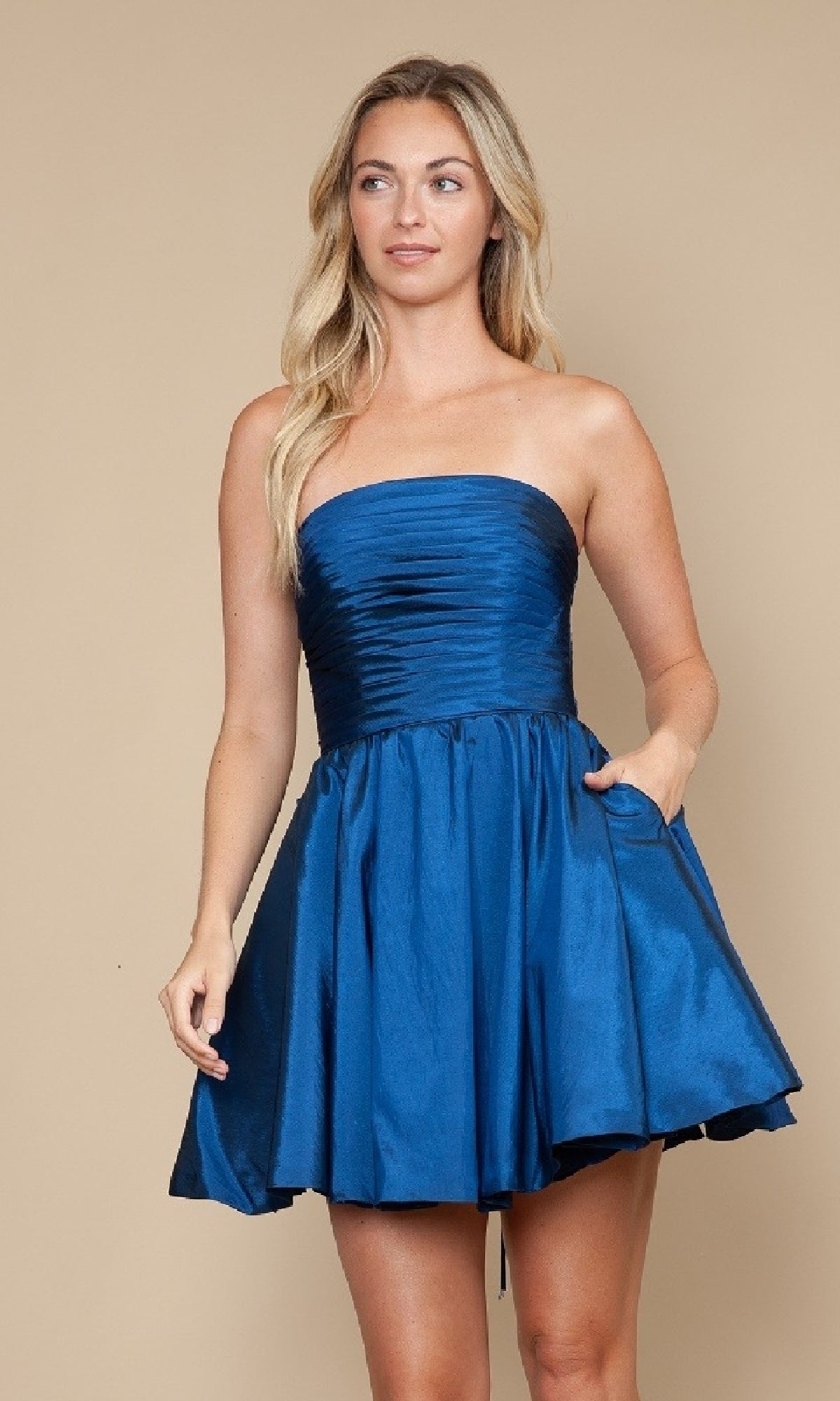 A woman wears the Strapless Short A-Line Hoco Dress: Poly USA 9908—a fitted blue satin mini with a flared skirt and side pocket—standing against a plain beige background.