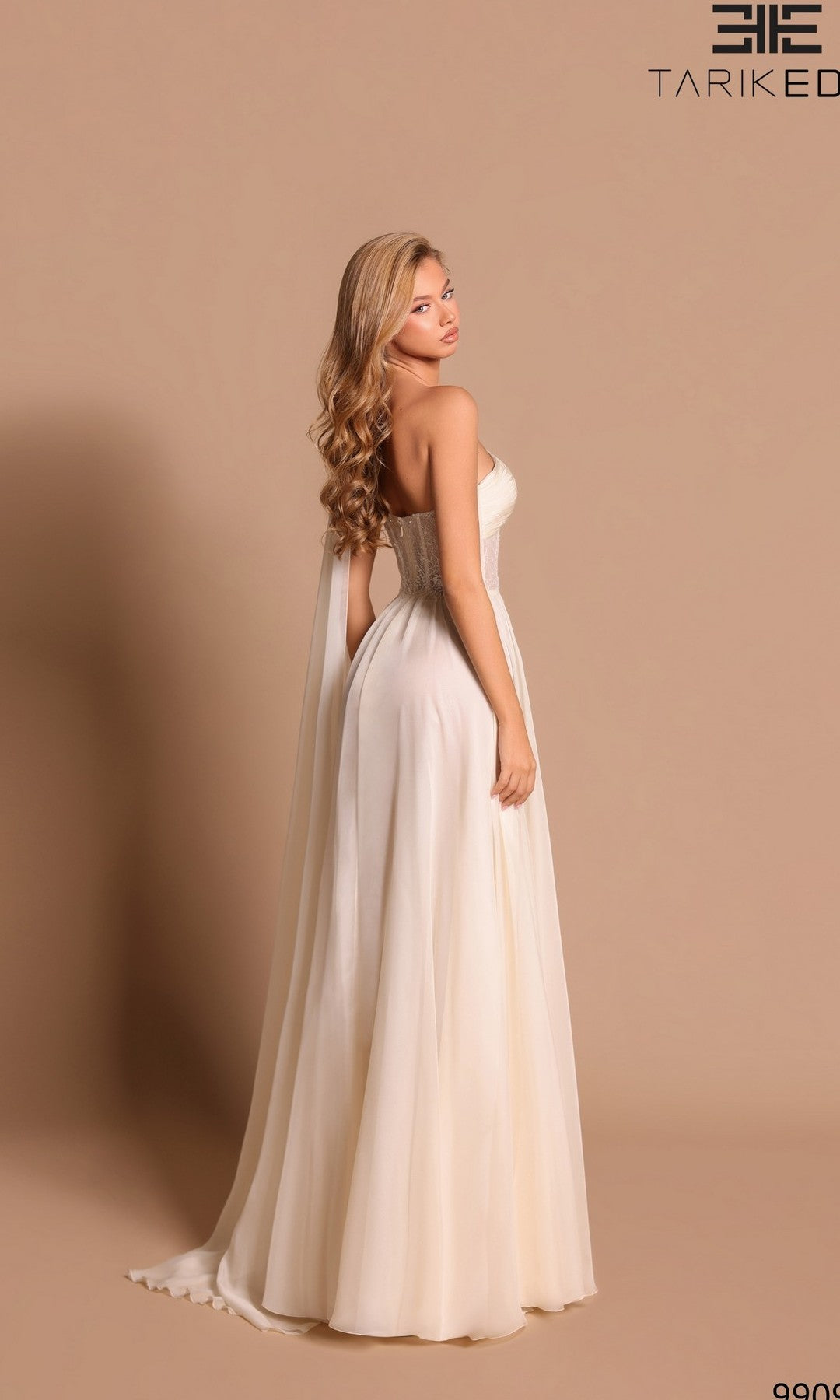 A woman in the Long Formal Dress: Tarik Ediz 99098, a strapless, floor-length ivory gown, stands against a beige background and looks over her shoulder.