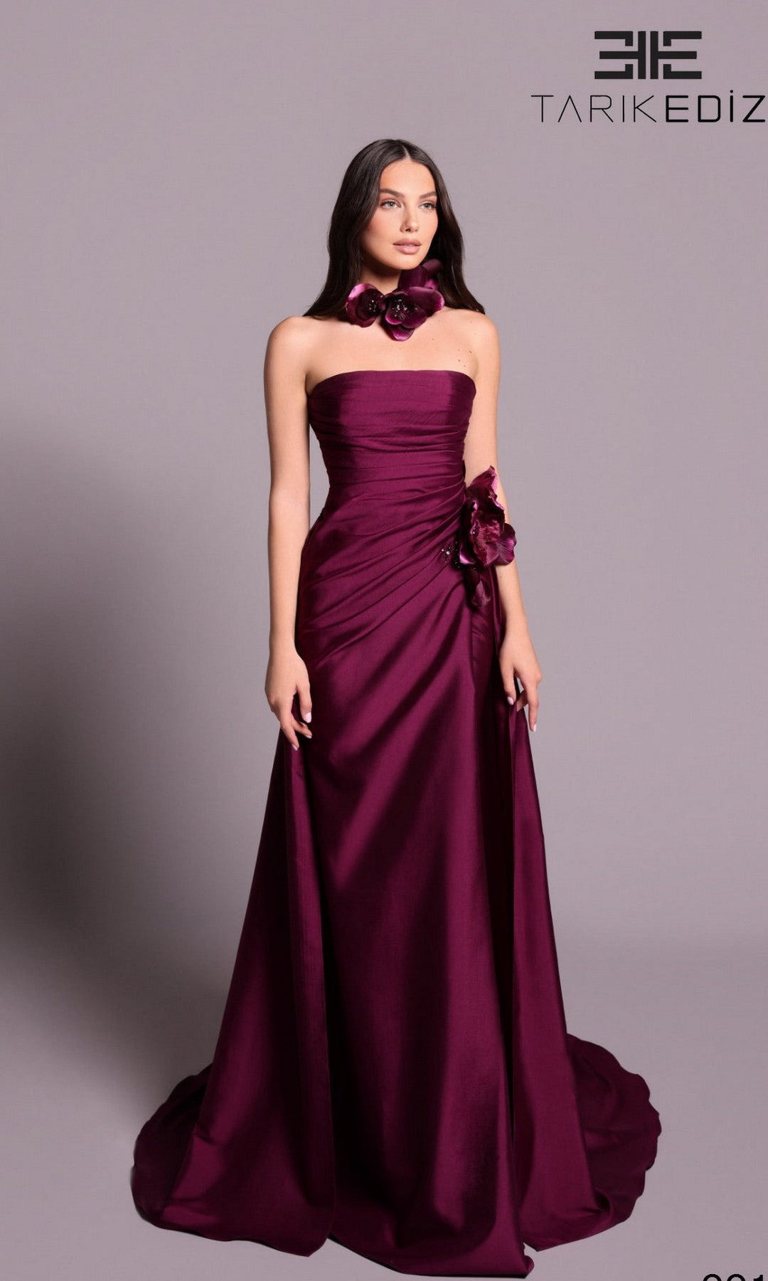 A woman stands against a plain background wearing the Tarik Ediz 99106 long formal dress in Cranberry Juice, a floor-length, strapless satin gown with floral accents, draped detailing, form-fitting silhouette, and matching neckpiece.