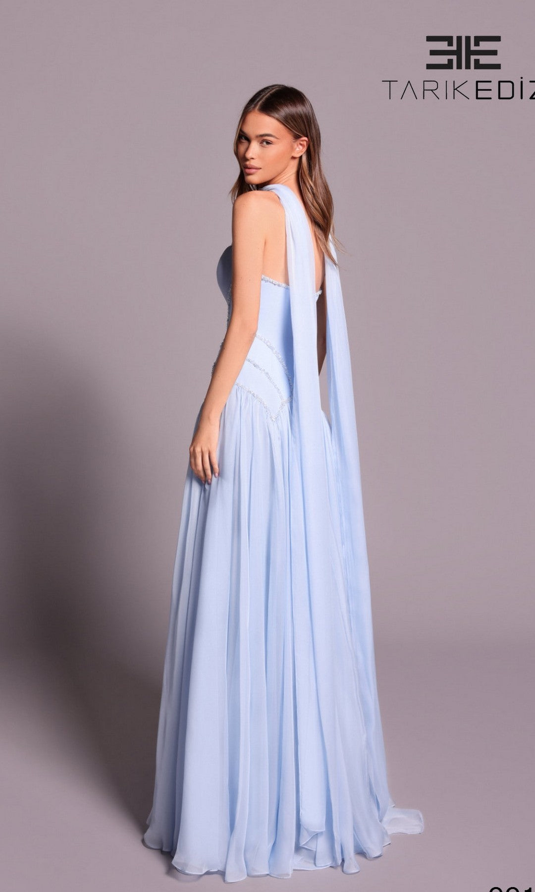 A woman poses in the Tarik Ediz 99136, a light blue, floor-length A-line chiffon dress with a draped scarf detail and a flowing skirt.