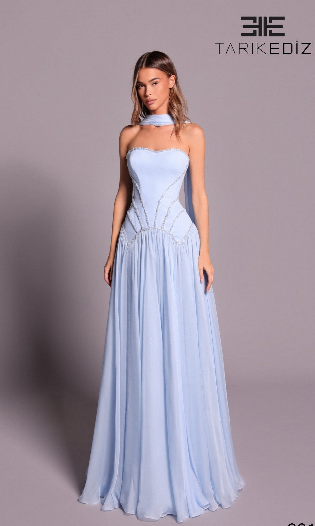 A woman in the Tarik Ediz 99136 Long Formal Dress stands against a plain background. The Blue Aura strapless chiffon gown features an embellished bodice, matching choker, floor-length A-line skirt, and a beautifully fitted silhouette.