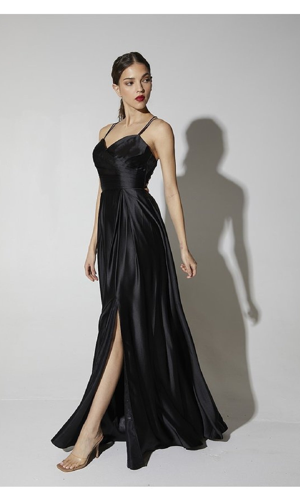 A woman wearing the Strappy-Back Long Satin Prom Dress Eureka 9925, a black gown with spaghetti straps and a thigh-high slit, stands against a plain white background.