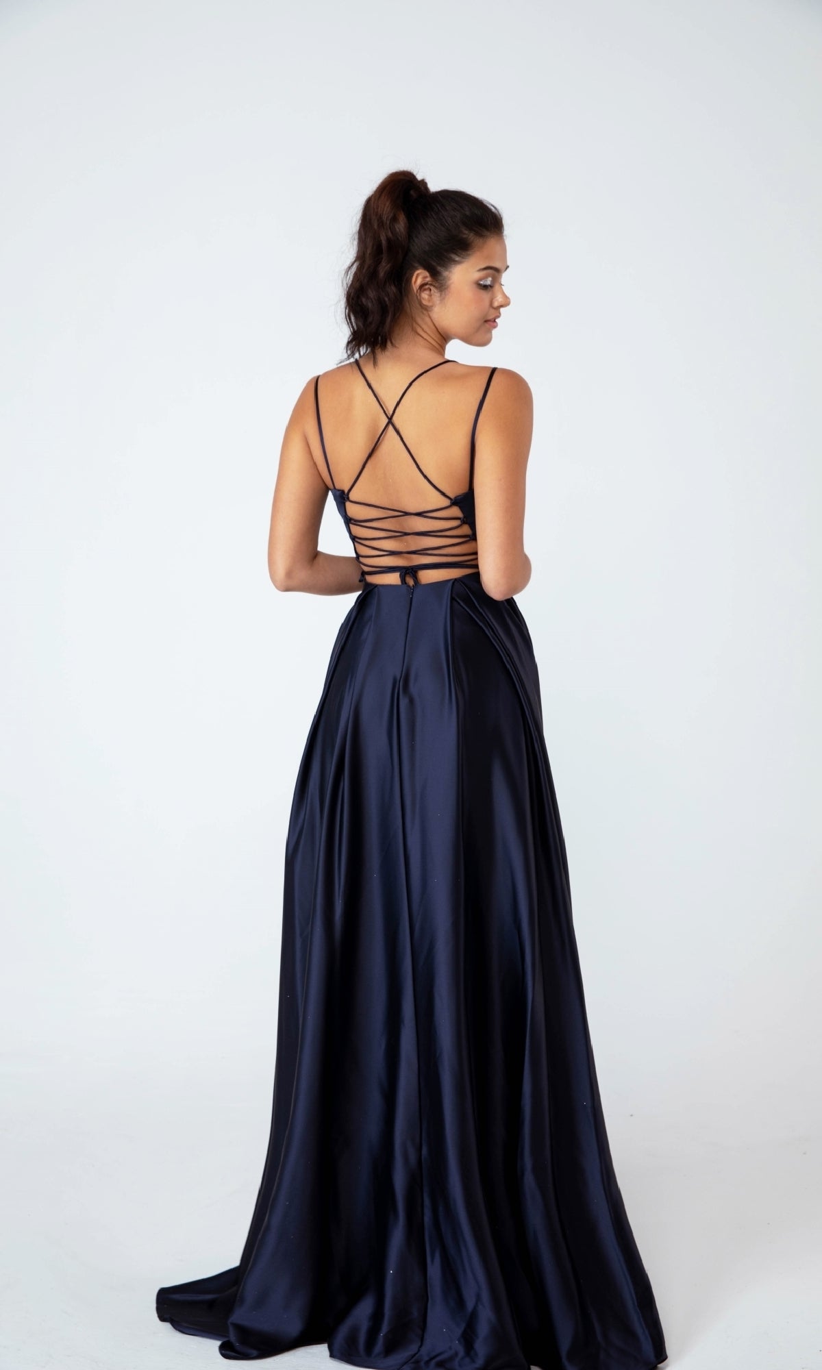 A woman stands facing away against a plain white background, modeling the Strappy-Back Long Satin Prom Dress Eureka 9925 in dark blue with a strappy open back and elegant A-line silhouette.