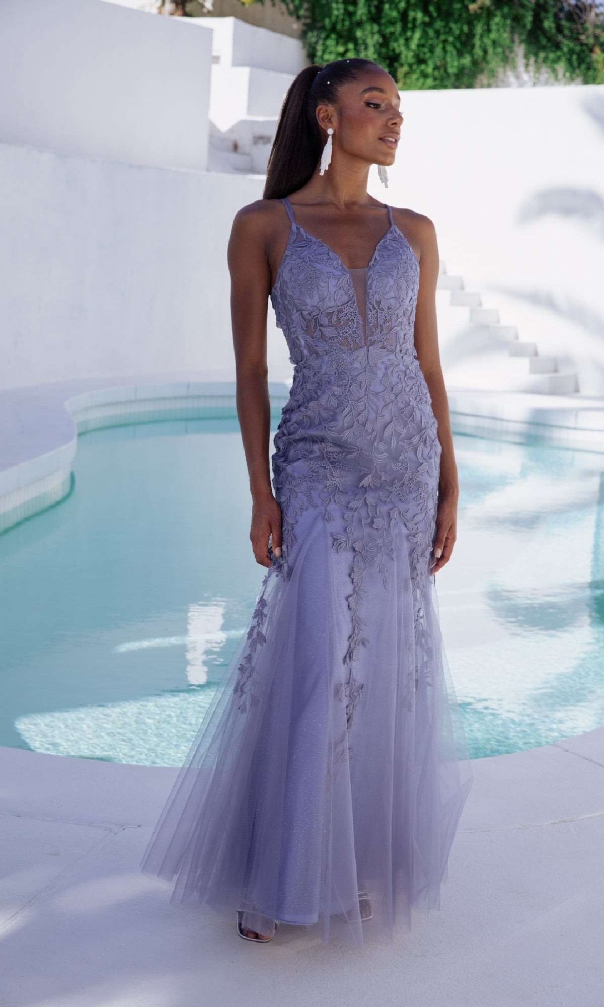 A woman stands by a pool outdoors wearing the Eureka 9958 Embroidered-Lace Long Prom Dress, a lavender gown with strappy back and floral embellishments.