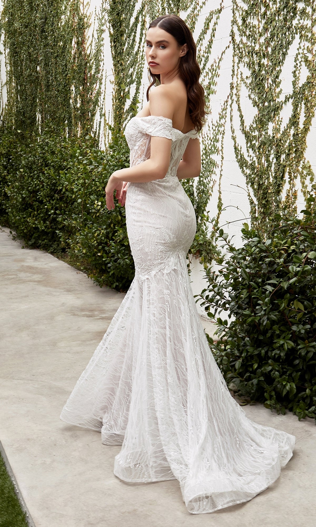 A woman in an Off-the-Shoulder Long White Lace Prom Gown with a mermaid silhouette and sweeping train stands on a walkway near green bushes and a vine-covered wall.