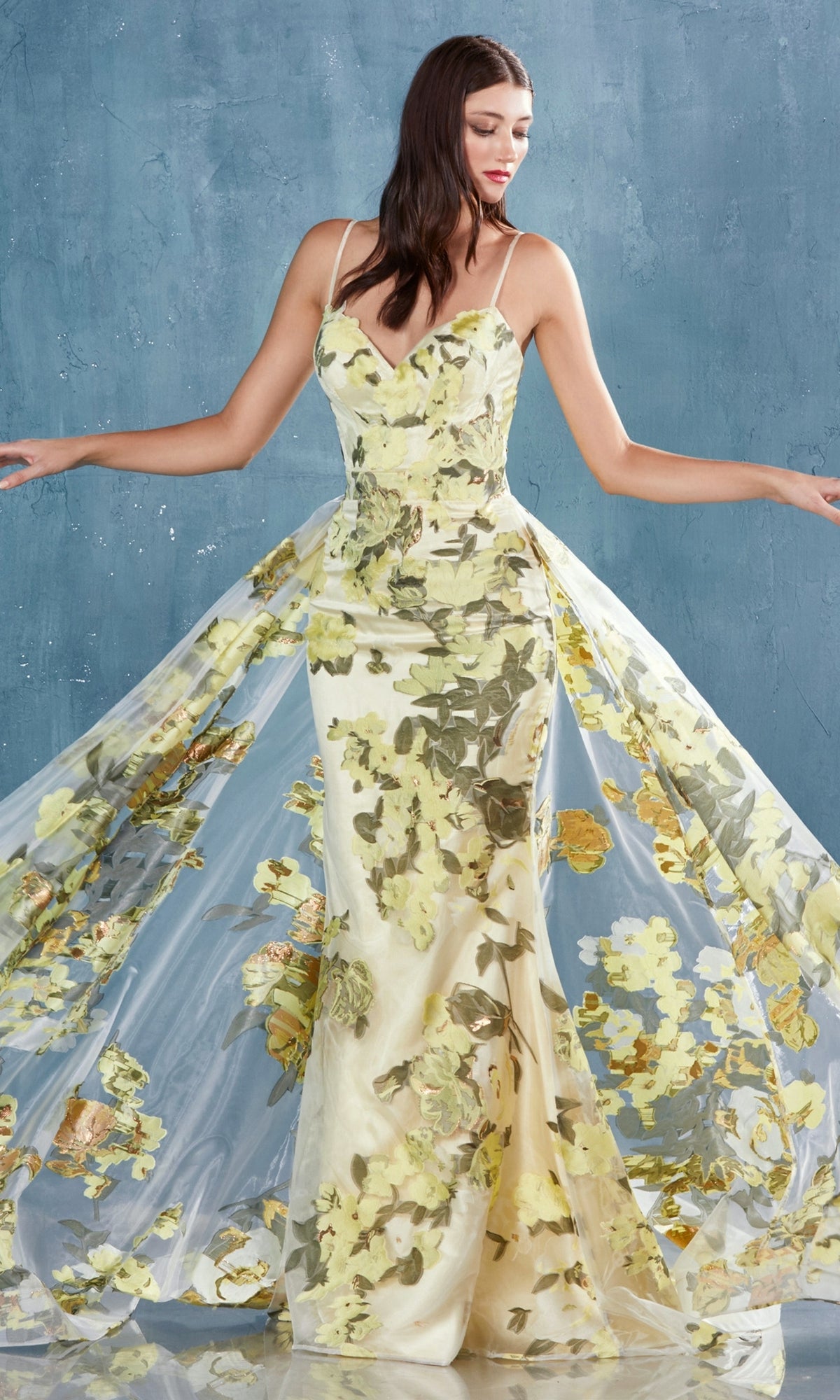A woman poses against a textured blue background wearing the Lemon Yellow Floral-Print Long Prom Dress A0770, featuring a fitted silhouette, thin straps, and a sheer flowing overlay.