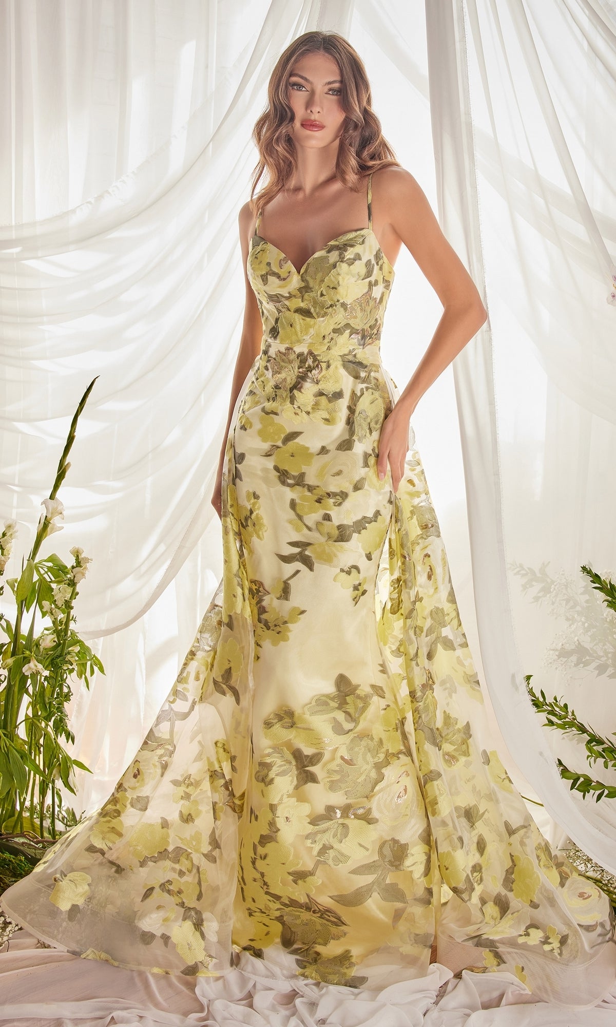 A woman stands before sheer curtains in the Lemon Yellow Floral-Print Long Prom Dress A0770, featuring spaghetti straps and a flowing skirt. Surrounded by lush green plants, she embodies a fresh, elegant vibe.