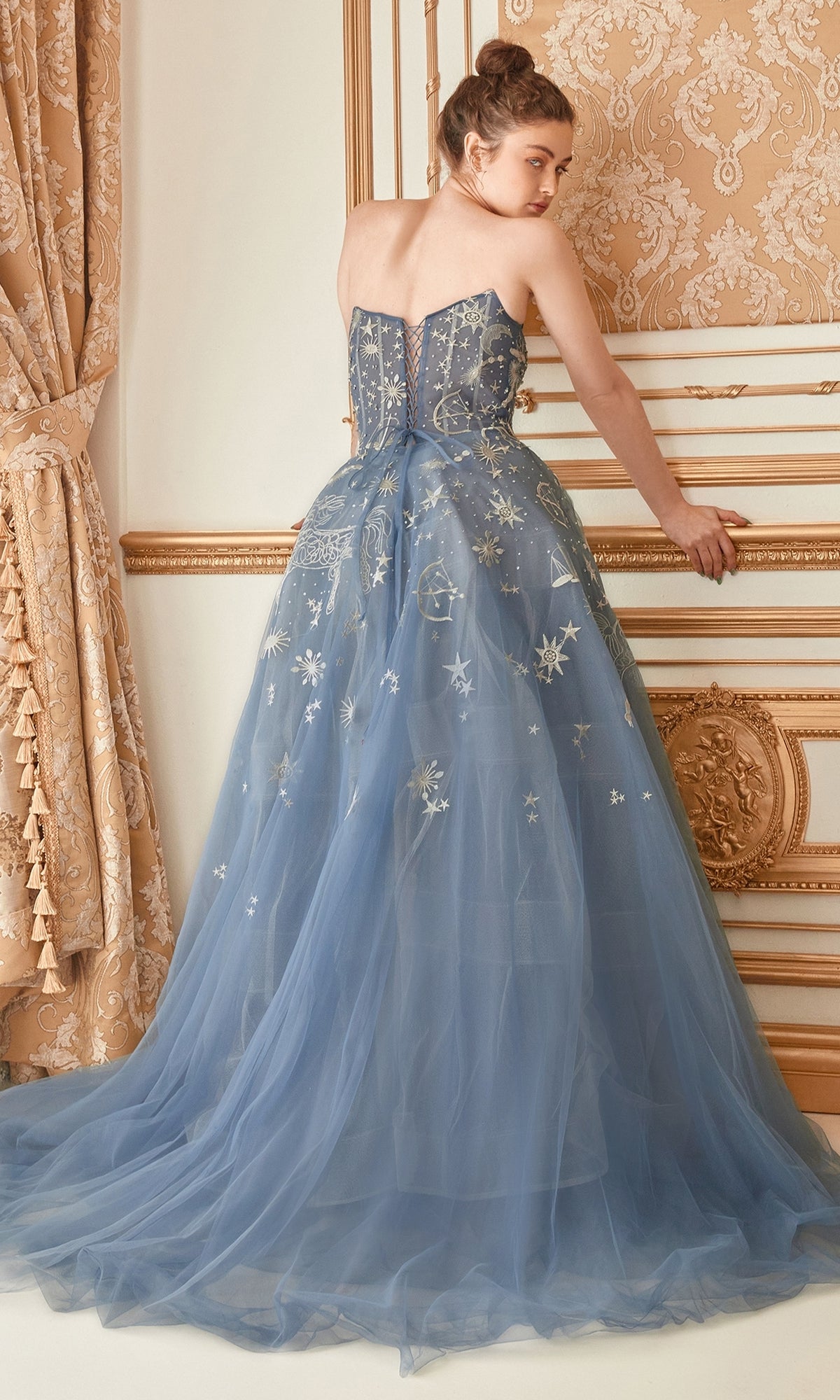 A woman faces away from the camera wearing the Constellation-Print Strapless Prom Ball Gown A0890, a floor-length blue dress with star motifs, in an ornate gold-accented room.
