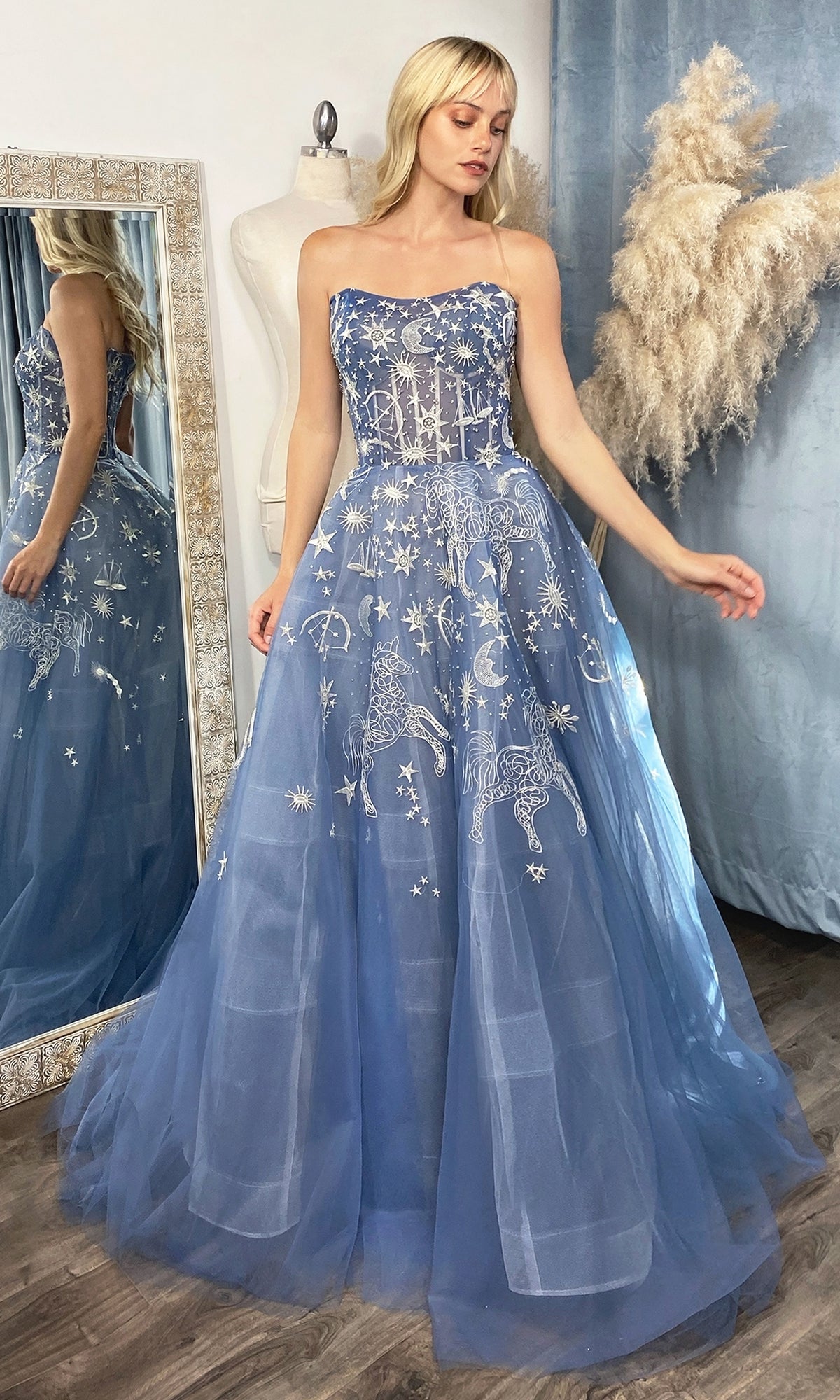 A woman poses by a mirror and pampas grass in a well-lit room with wooden floors and blue curtains, wearing the Constellation-Print Strapless Prom Ball Gown A0890 featuring blue fabric and silver celestial embroidery.