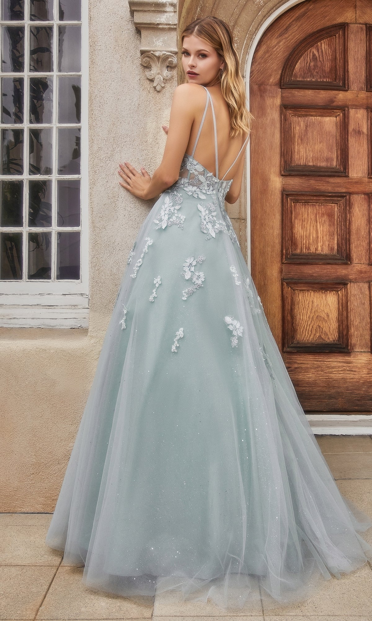 A woman in a sage green, floor-length glitter prom dress (style A0892) with delicate straps and floral appliqués stands facing a wooden door, glancing over her shoulder with timeless elegance.