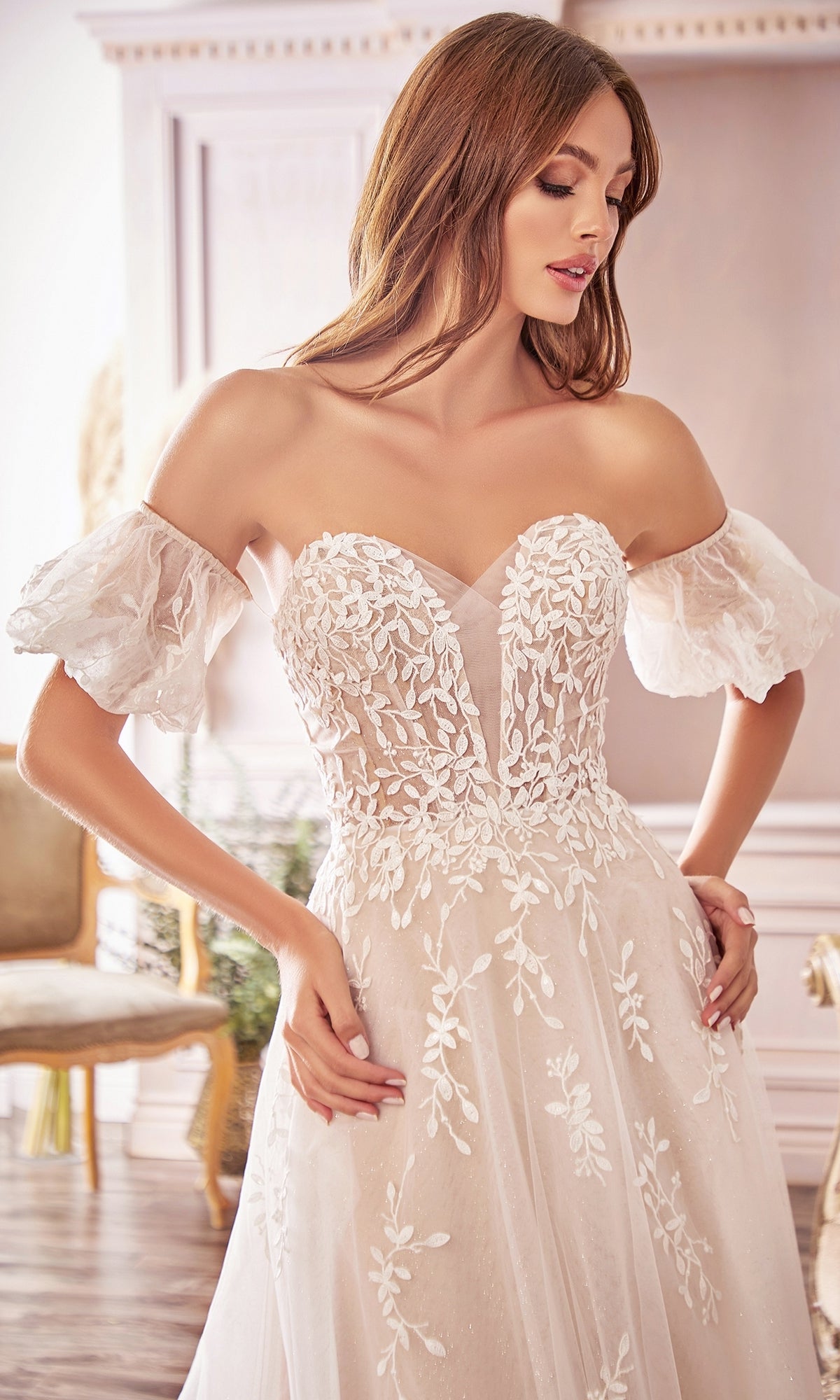 A woman in the Puff-Sleeve Long White Wedding Dress A1014, featuring floral lace and removable puff sleeves, stands indoors in a softly lit, elegant room.