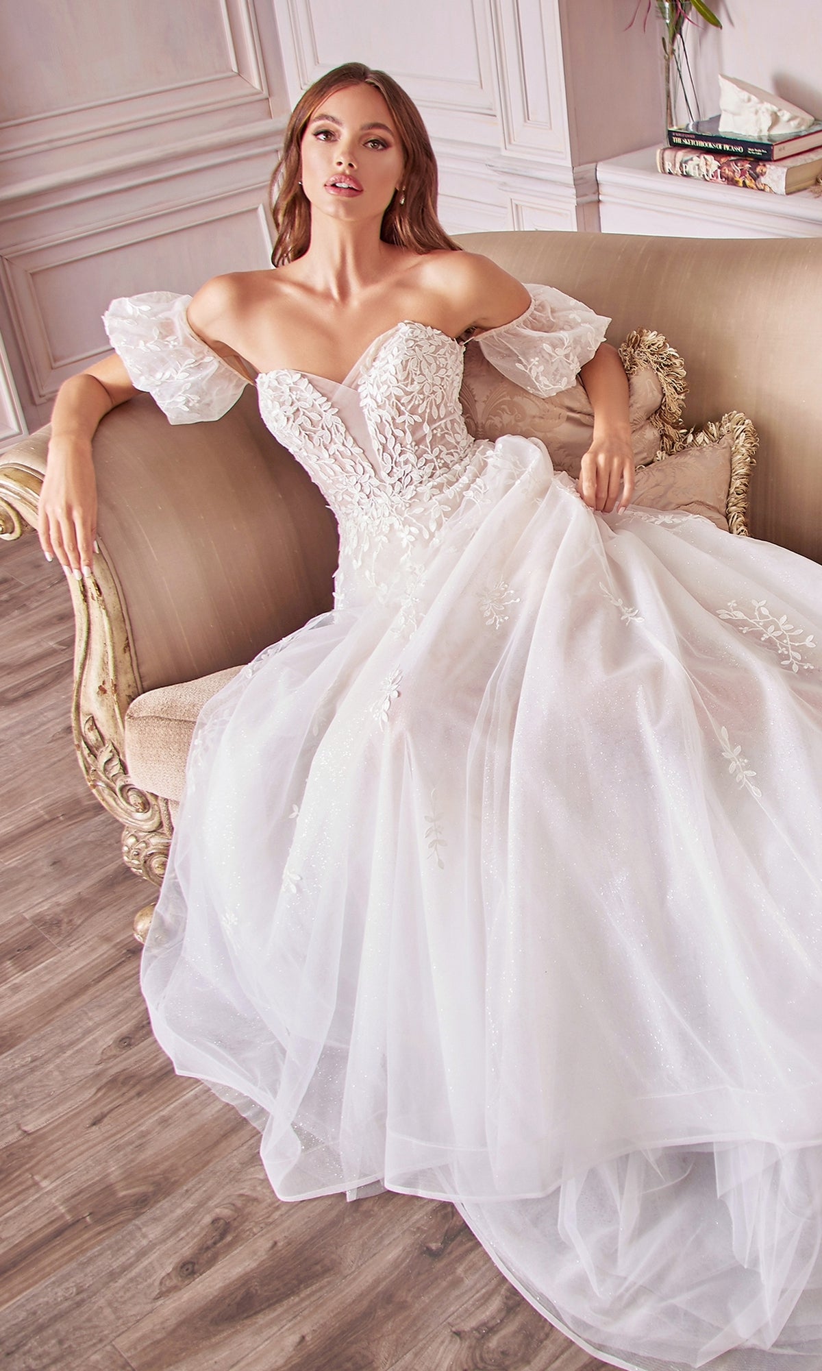 A woman in the Puff-Sleeve Long White Wedding Dress A1014 with off-the-shoulder lace, removable puff sleeves, and a full tulle skirt sits on a beige sofa in an elegant room with wood floors.