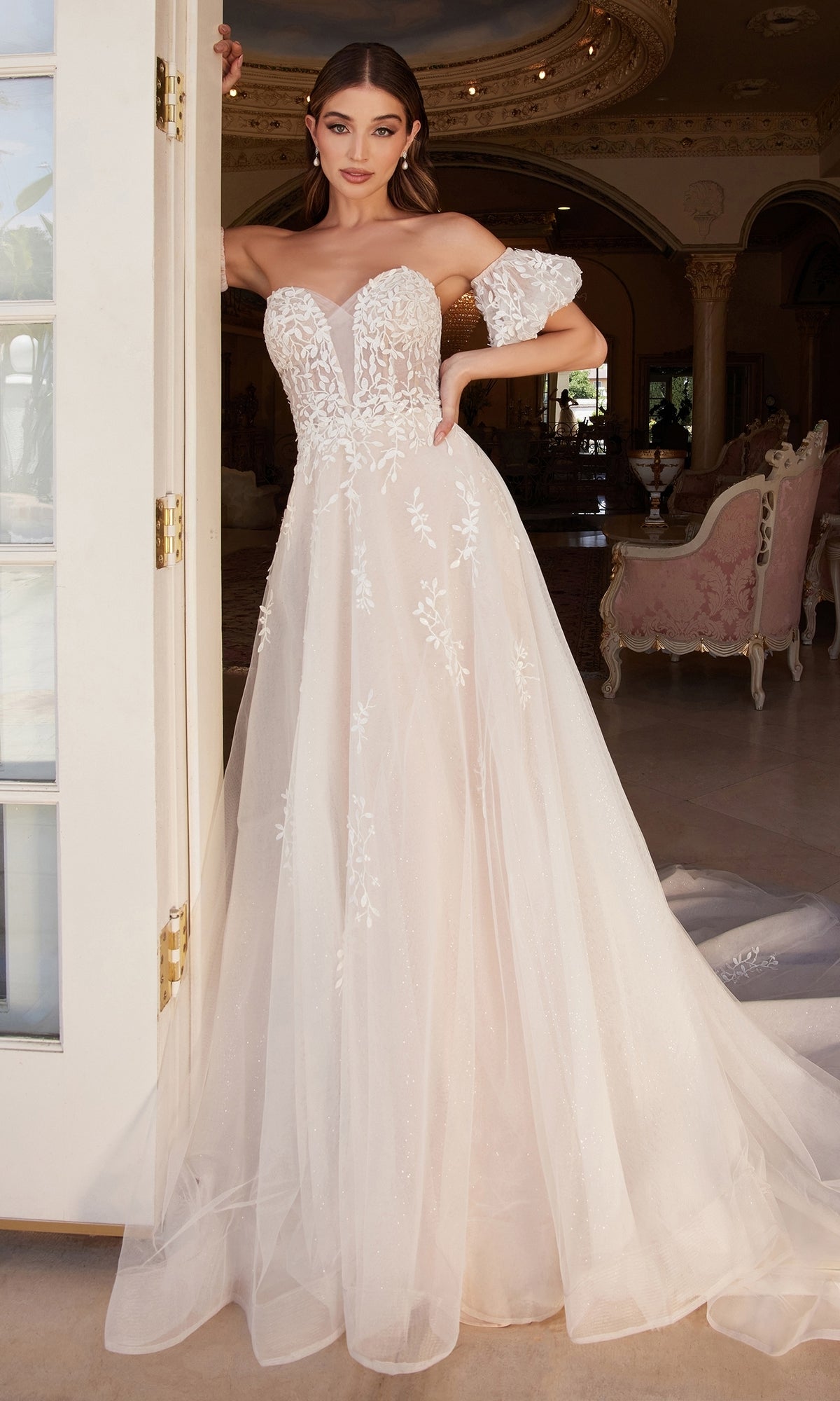 A woman stands in a doorway, wearing the Puff-Sleeve Long White Wedding Dress A1014—strapless with floral lace details—holding one removable puff sleeve, elegant furniture visible in the background.
