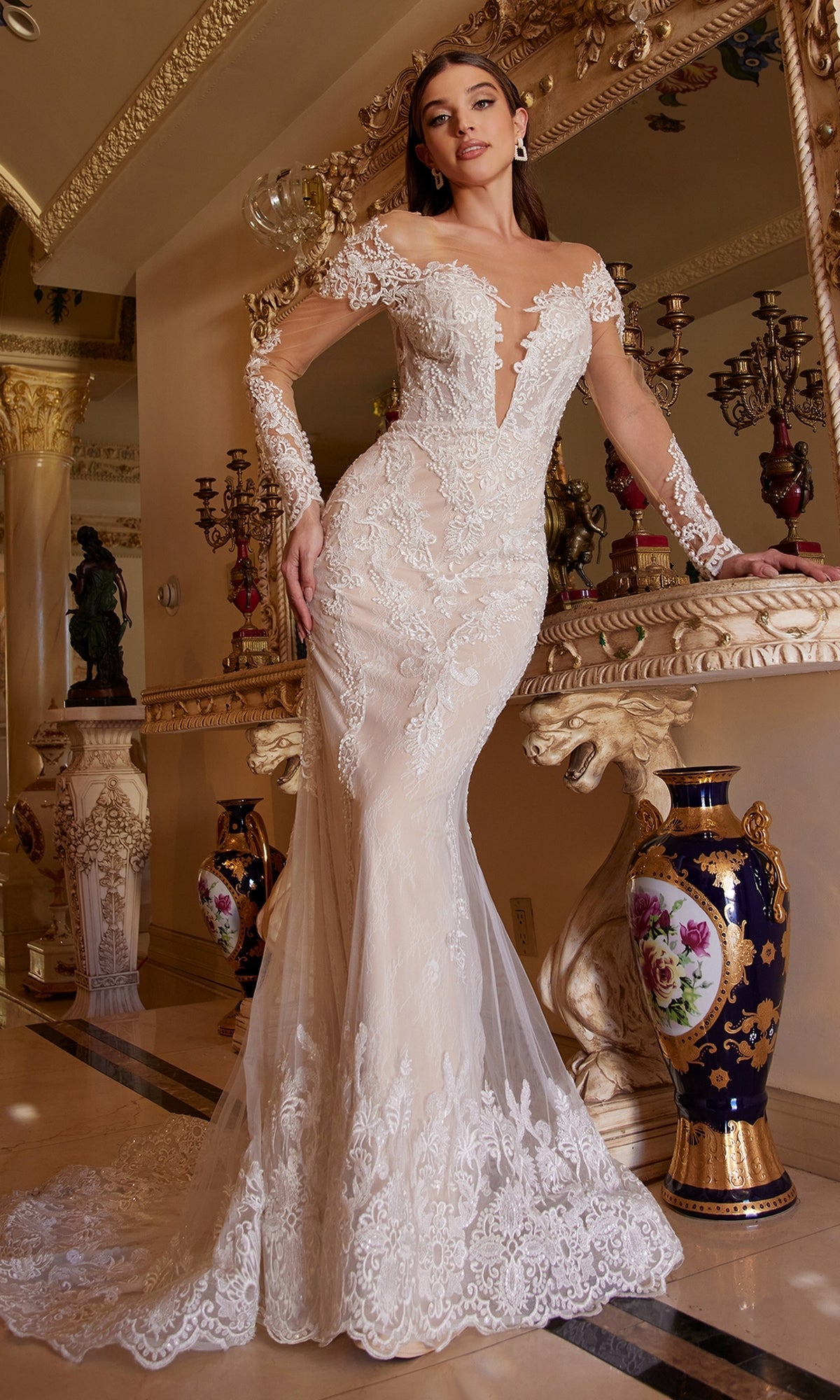 Wearing the Ladivine A1022 long white lace wedding dress, a woman stands by an ornate mirror in a lavish, gold-accented room decorated with floral vases and statues.