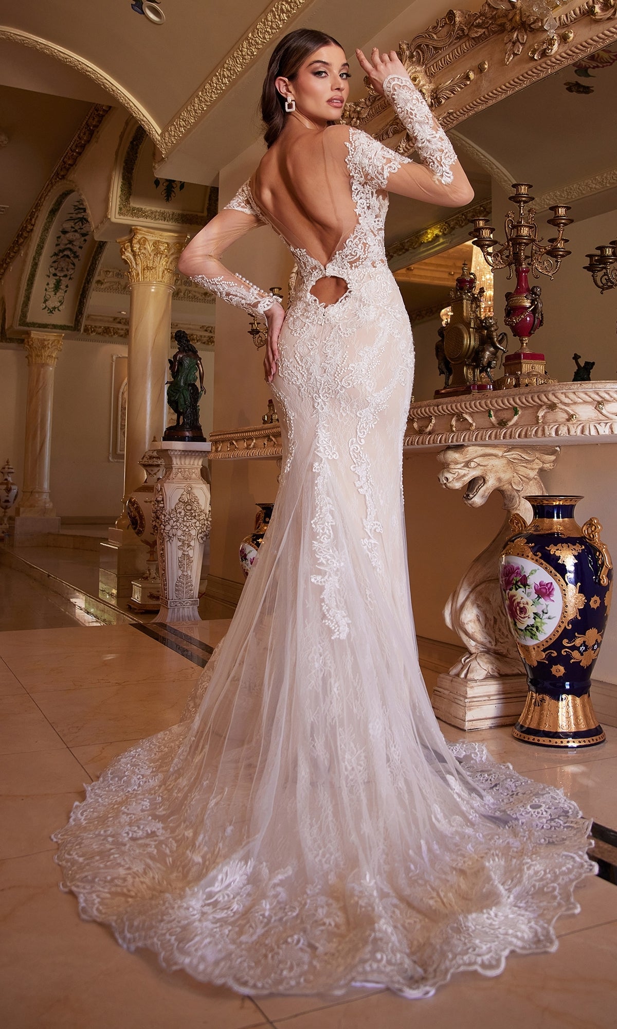 A woman poses indoors in the Ladivine A1022, a long white lace wedding dress with an open back and long sleeves, standing on a polished floor near ornate decor and columns.