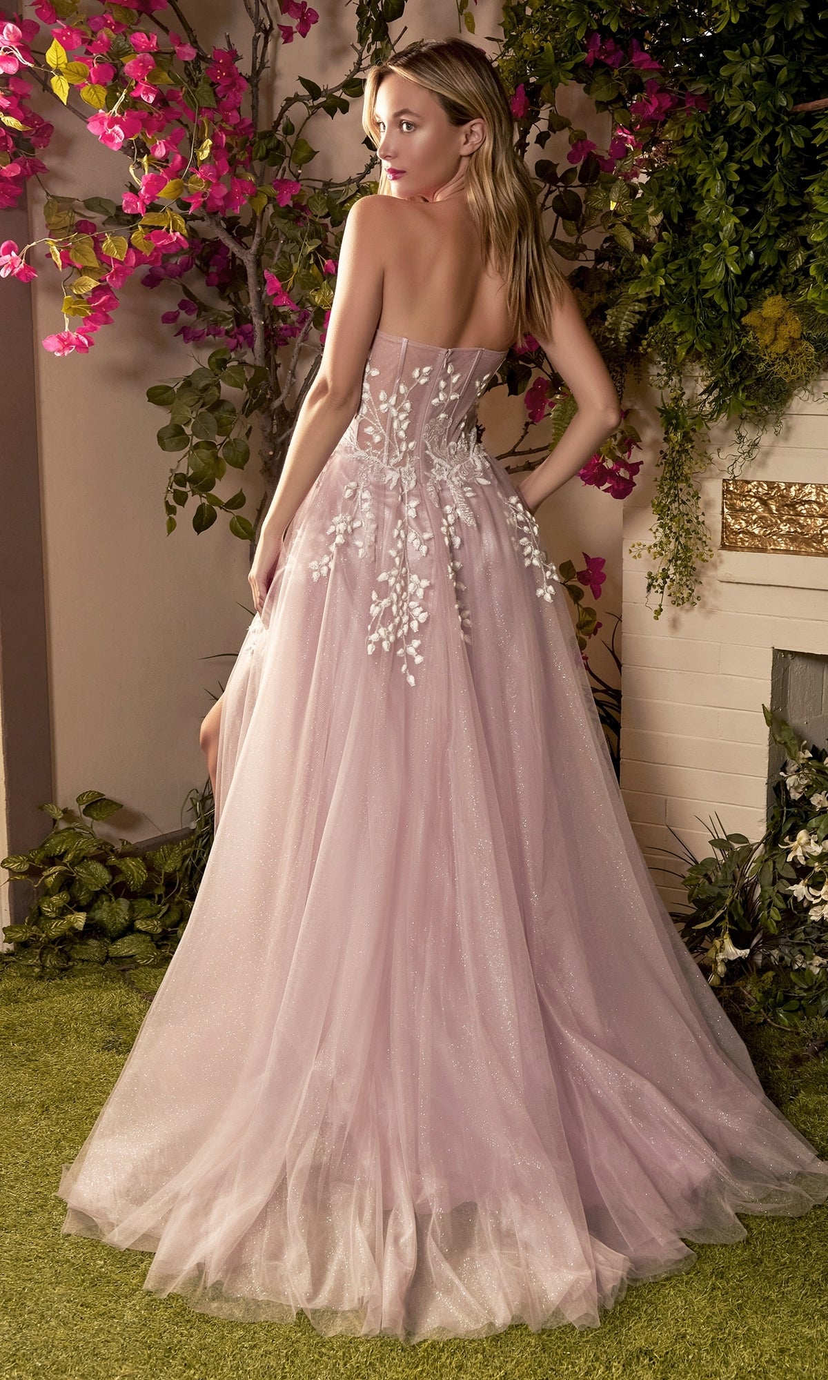 A woman stands with her back to the camera in a light pink Strapless Sweetheart Prom Ball Gown A1029 with floral embroidery, surrounded by lush greenery and pink flowers in a garden setting.