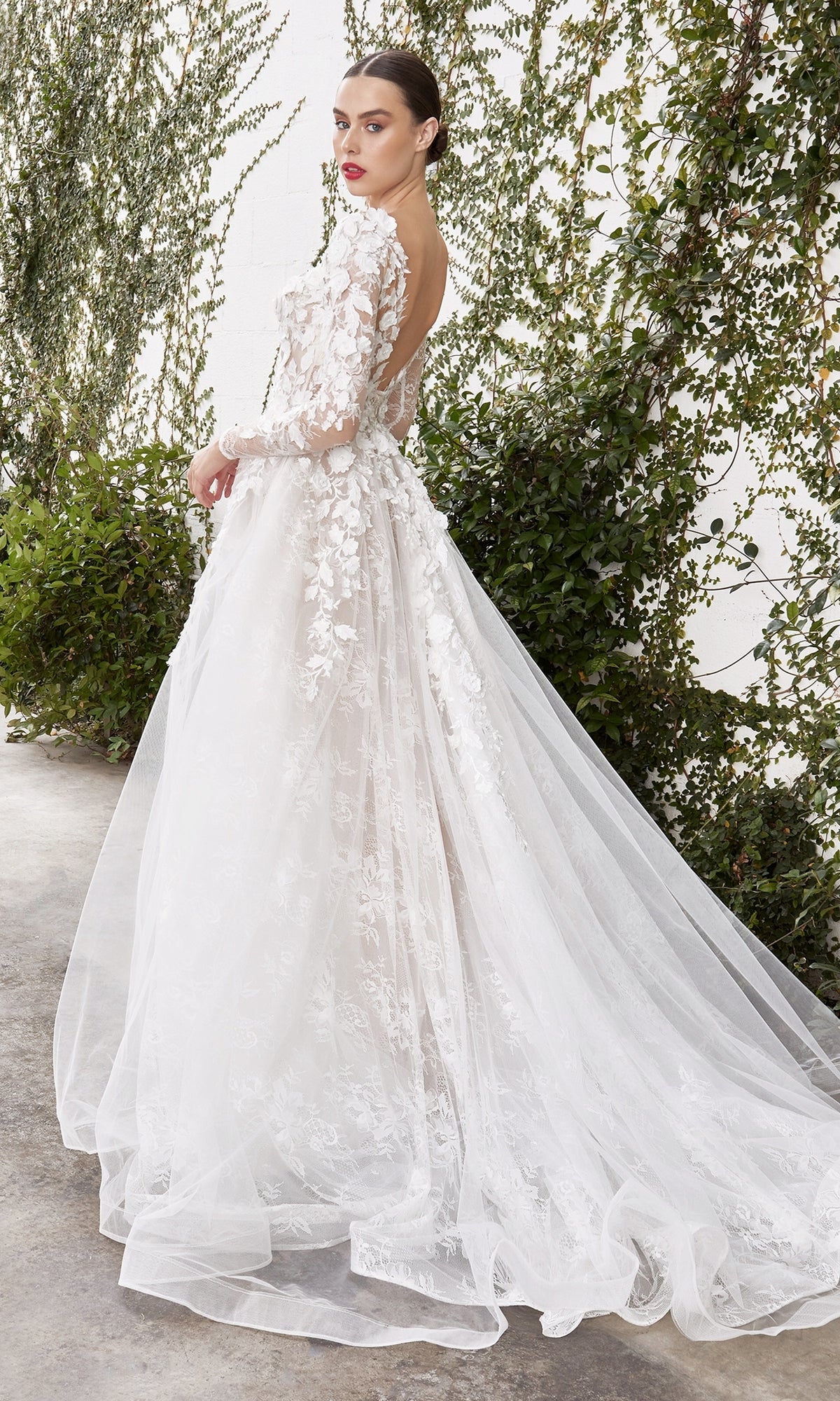 A woman in the Long Sleeve White Lace Bridal Gown: LadivineA1067W with a long train stands outdoors by a green vine-covered wall.