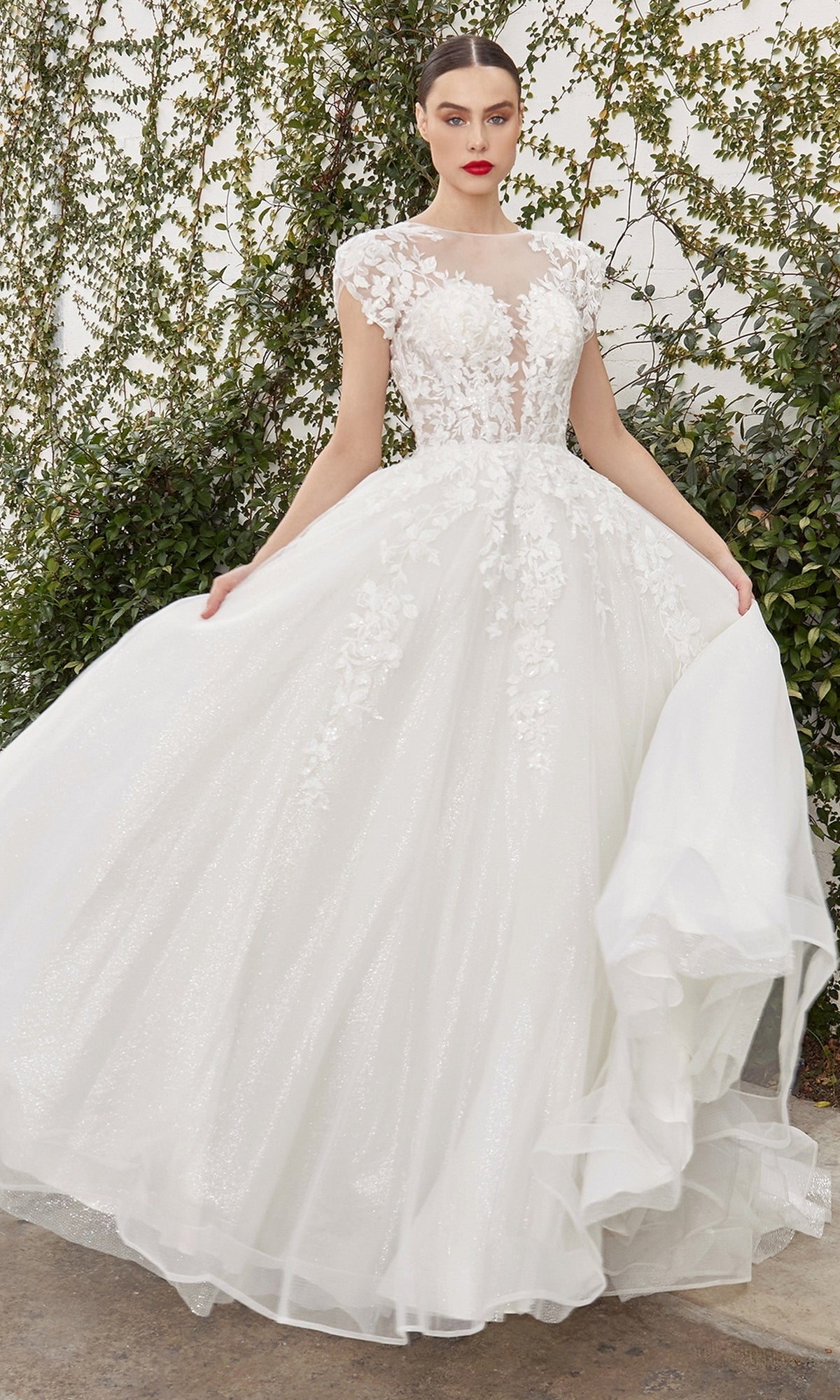 A woman in the Cap-Sleeve White Wedding Gown: Ladivine A1082W, featuring floral lace and a sheer neckline, stands outside beside a vine-covered wall.