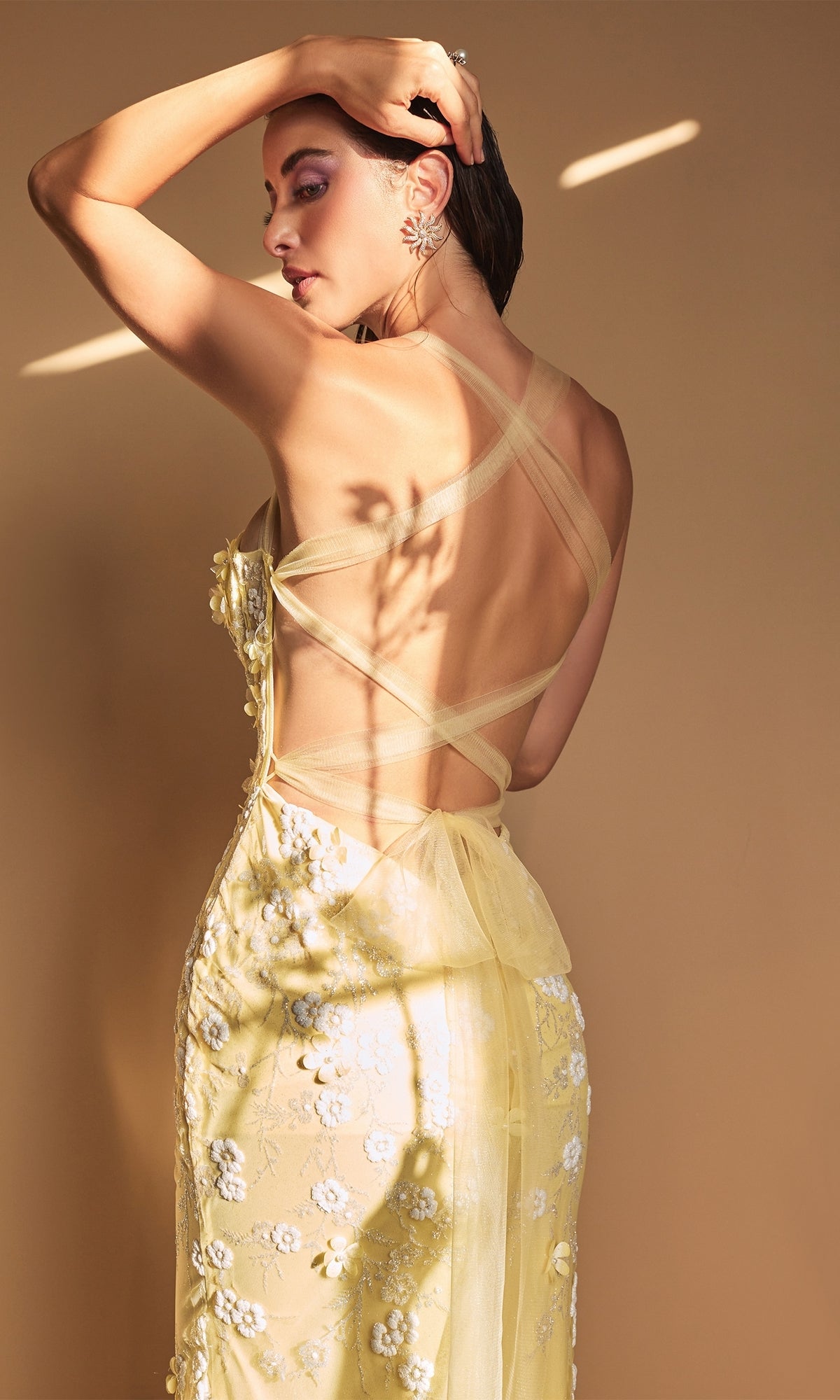 A woman in the Long Formal Mermaid Prom Dress with Daisies A1115, featuring yellow floral embroidery and a sheer crisscross back, stands against a beige background in partial natural light.