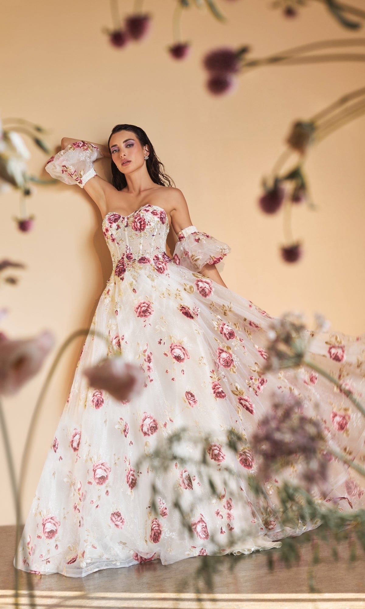 Woman wearing the Puff-Sleeve Strapless Floral-Print Ball Gown A1133, featuring pink patterns and detached sleeves, stands against a beige background with blurred flowers in the foreground.