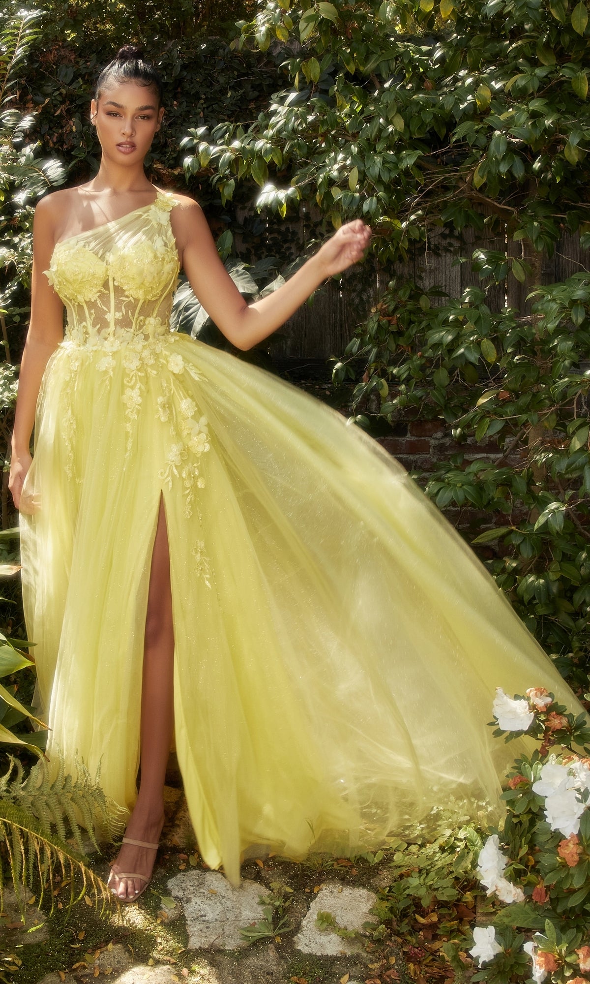 A woman stands outdoors in lush greenery and flowers, wearing the Sheer-Bodice One-Shoulder Prom Ball Gown A1140 in light yellow with floral details, a sheer corset bodice, and a high slit.