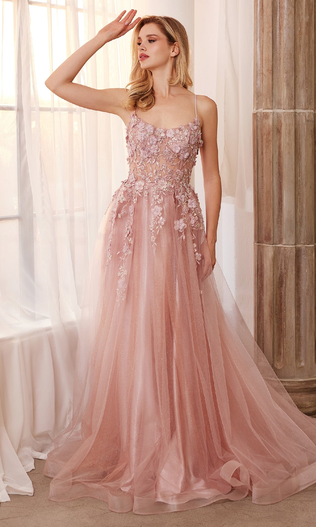 A woman stands by a window wearing the Flower-Embellished Long Tulle Prom Ball Gown A1142, a floor-length blush pink dress with 3D floral details and delicate sheer accents.