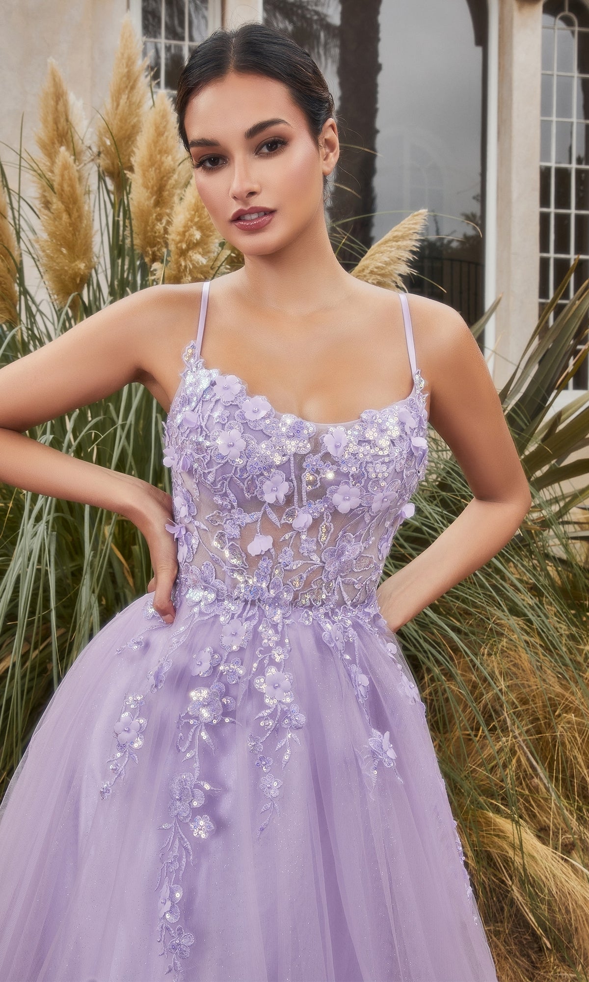 A woman stands outdoors in front of ornamental grasses and arched windows wearing the Flower-Embellished Long Tulle Prom Ball Gown A1142, featuring lavender tulle, 3D floral embellishments, and a sheer waist.