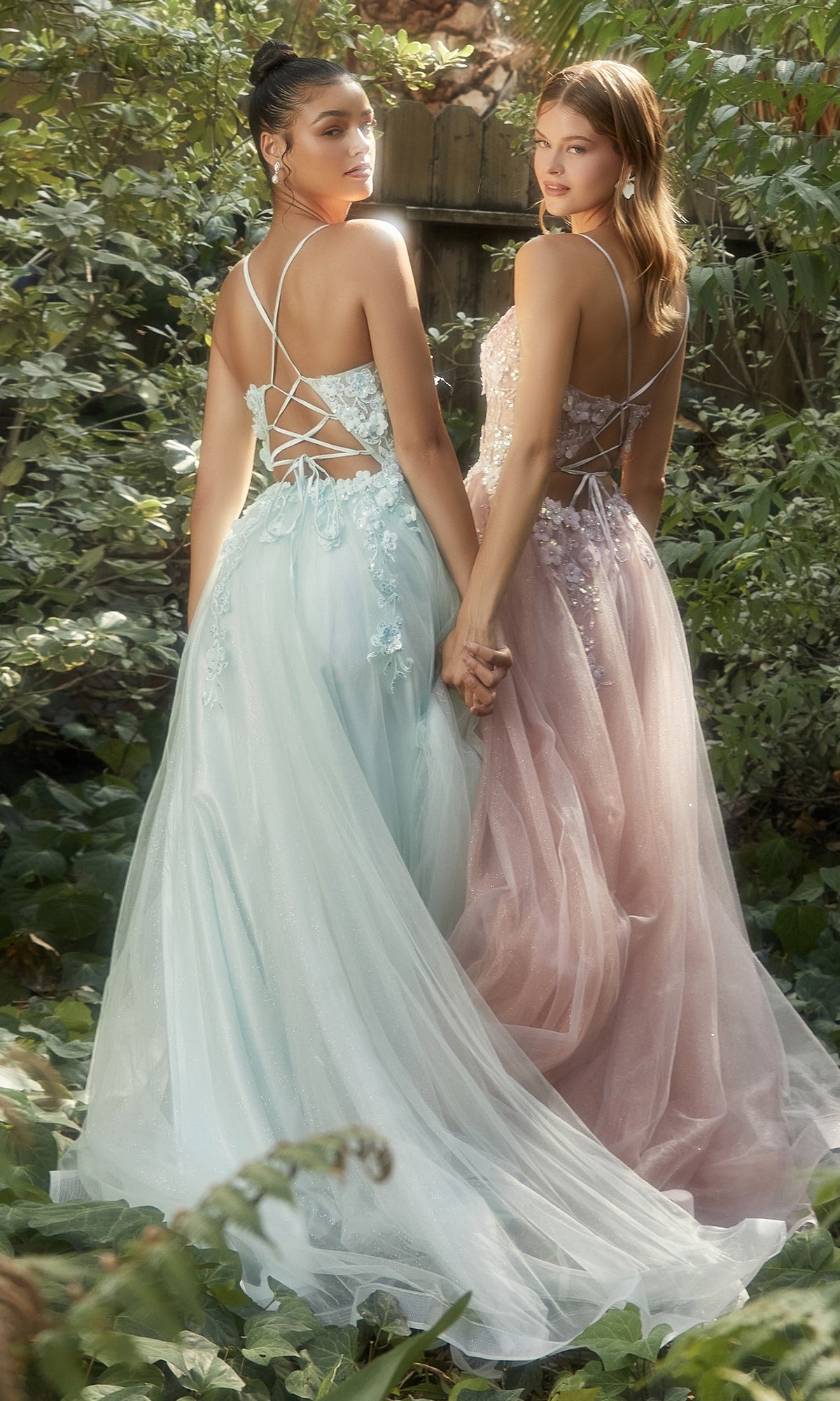 Two women stand side by side in a garden, holding hands and looking over their shoulders while wearing the Flower-Embellished Long Tulle Prom Ball Gown A1142, which features lace-up backs and subtle 3D floral accents.