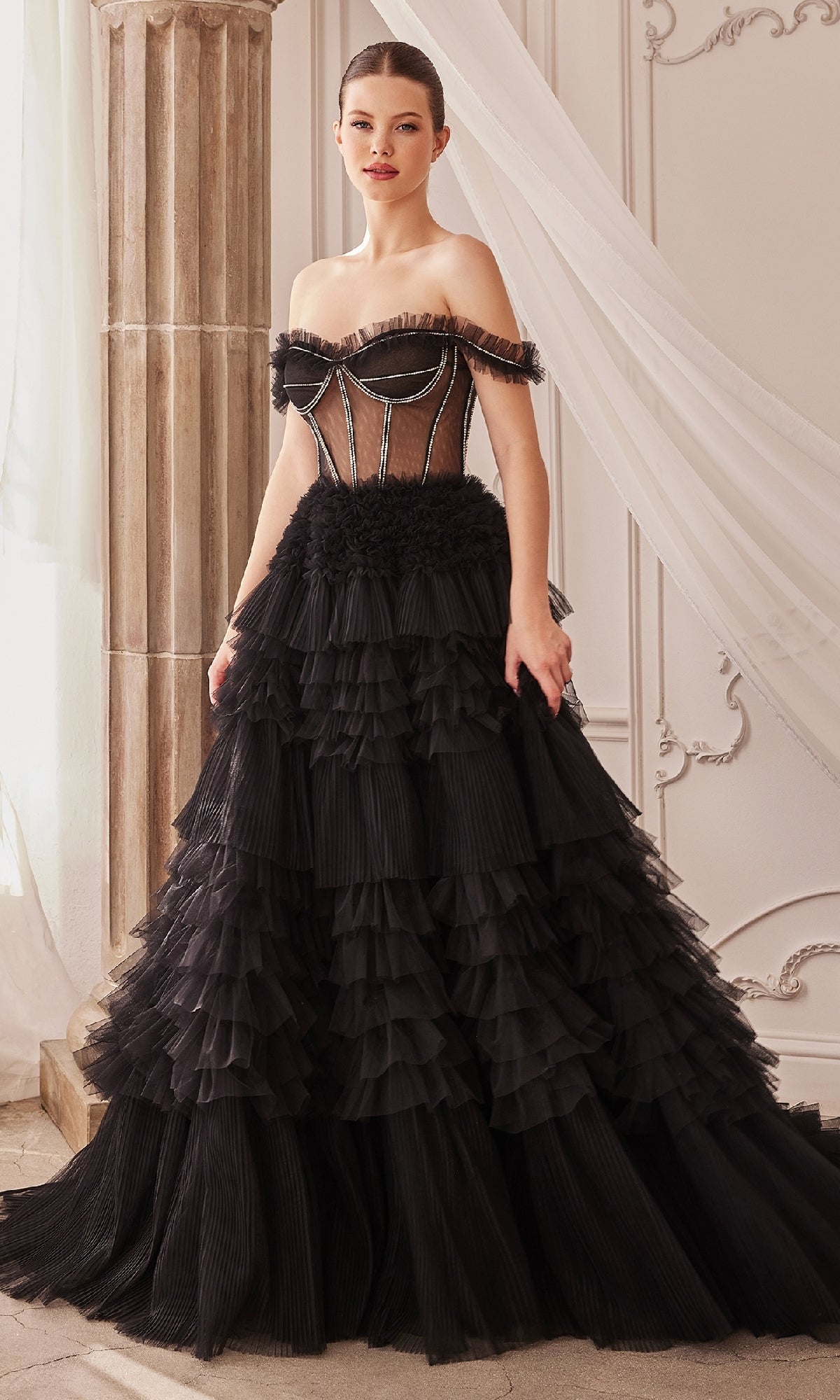 A woman indoors wears the Pastel Off-the-Shoulder Ruffled Ball Gown A1150, featuring ruffles, a sheer corset bodice, and a tiered skirt.