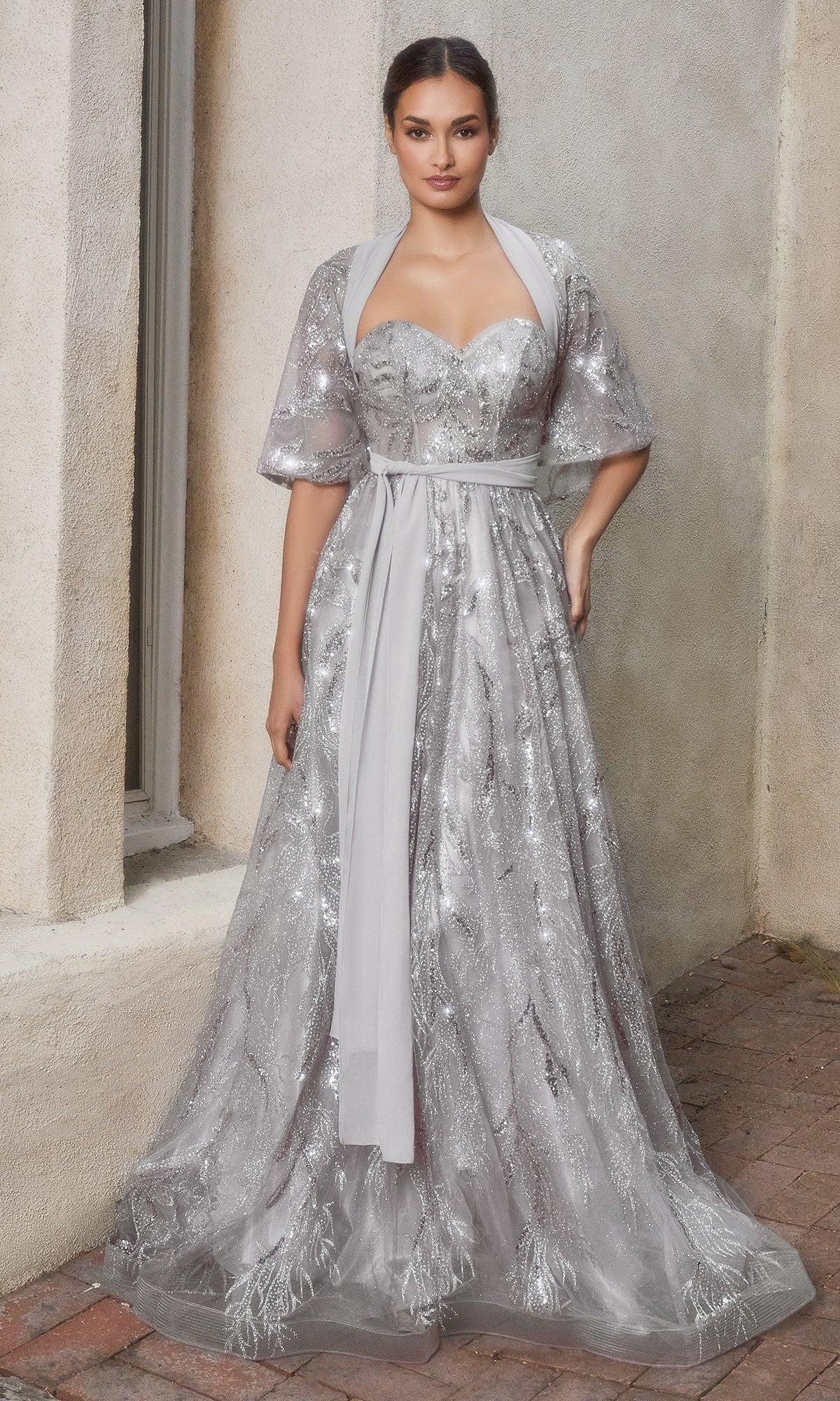 A woman stands against a textured wall wearing the Strapless Long Prom Ball Gown with Capelet A1174—a floor-length, silver sequined gown featuring a capelet and elegant detailing.