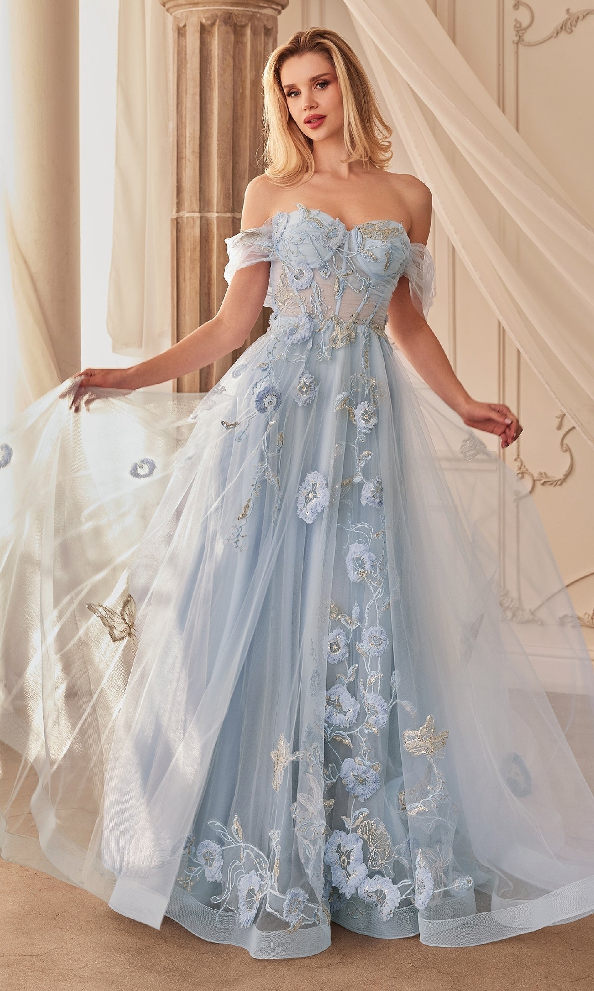 A woman wearing the Andrea and Leo A1246 Long Blue Prom Ball Gown with floral embroidery and sheer tulle stands indoors near classical columns and draped curtains.