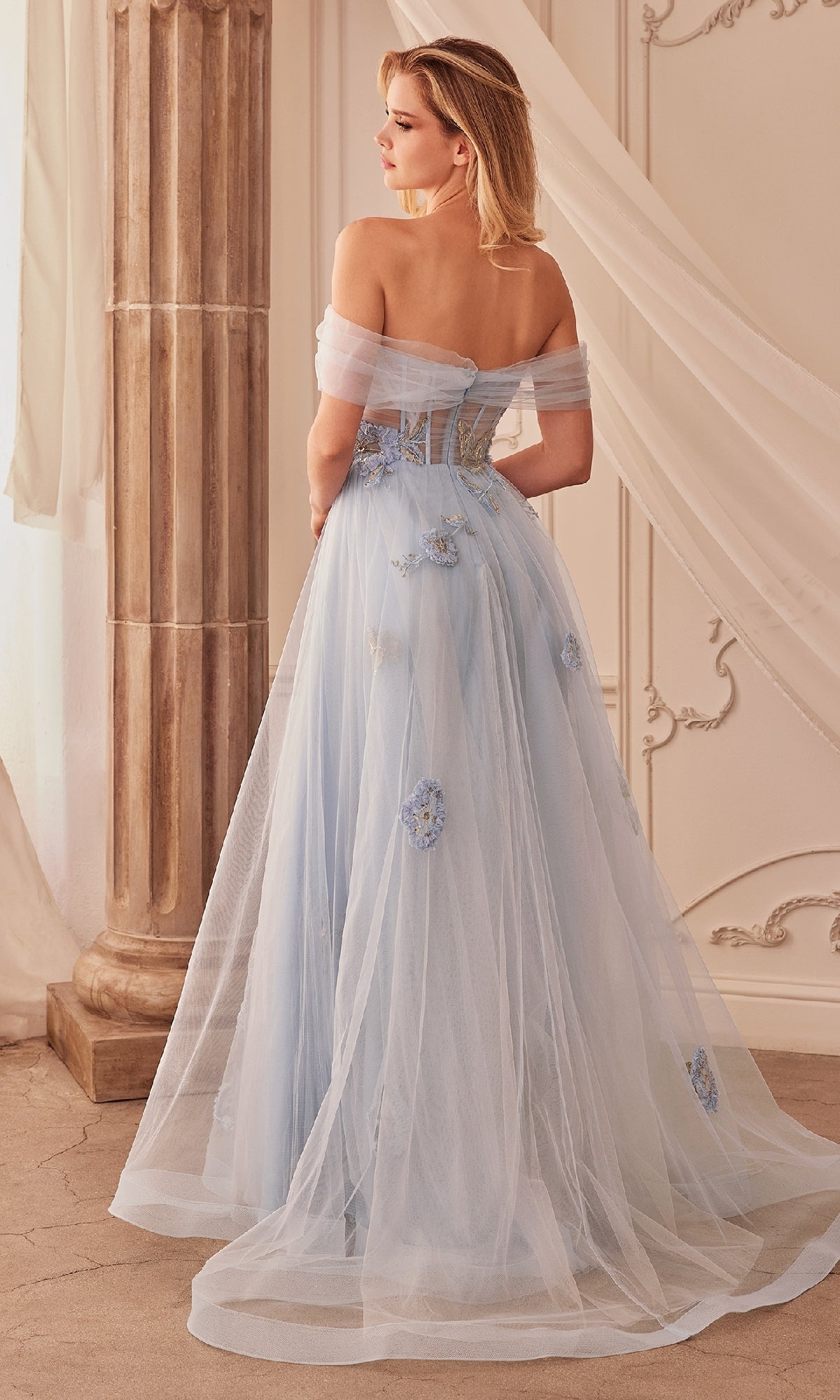 A woman in the Andrea and Leo A1246 Long Blue Prom Ball Gown stands with her back to the camera near a column, highlighting the ethereal elegance and floral embroidery of her formal evening dress.