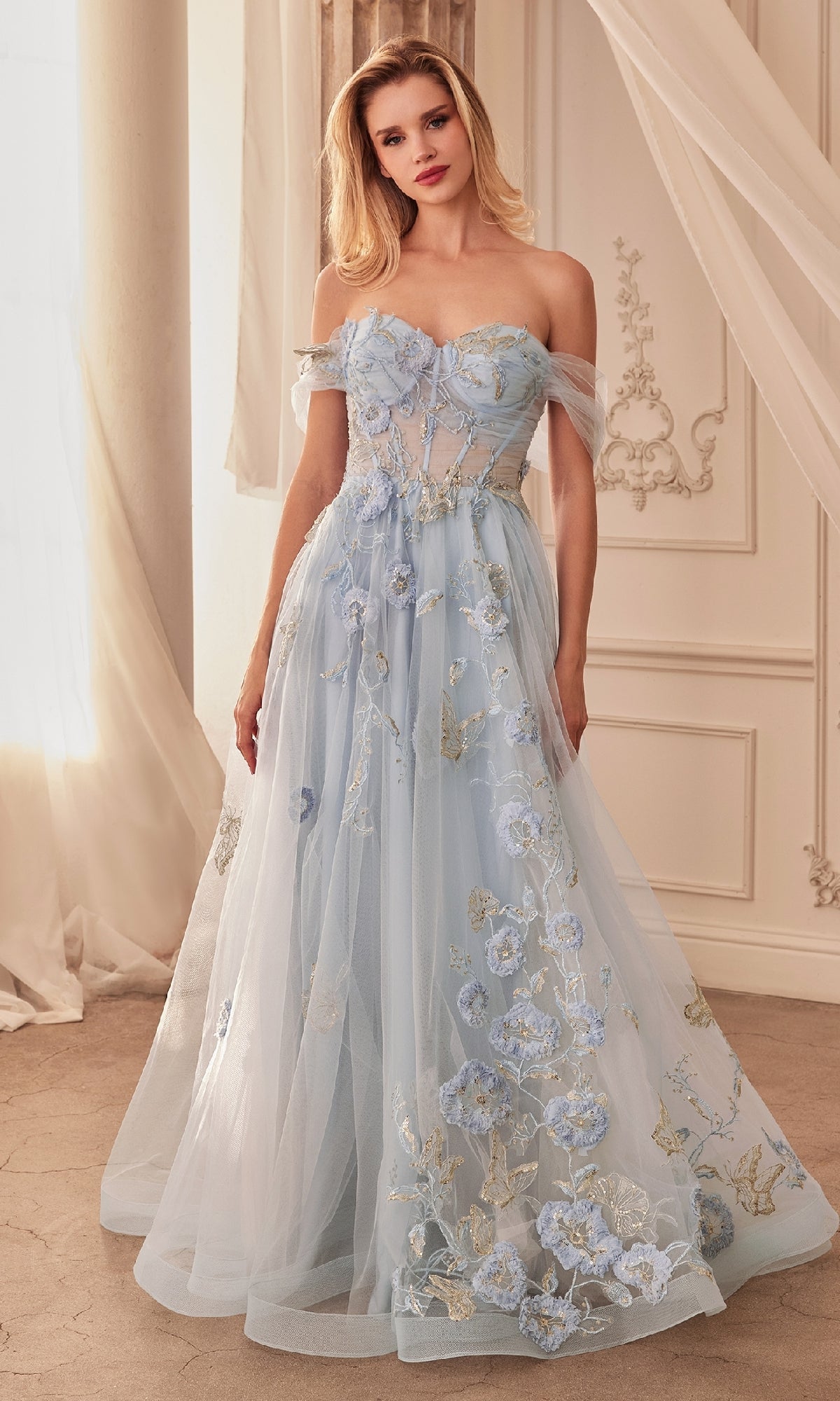 A woman stands indoors wearing the Andrea and Leo A1246 Long Blue Prom Ball Gown, styled off-the-shoulder with a floor-length silhouette, floral embroidery, and sheer tulle details.