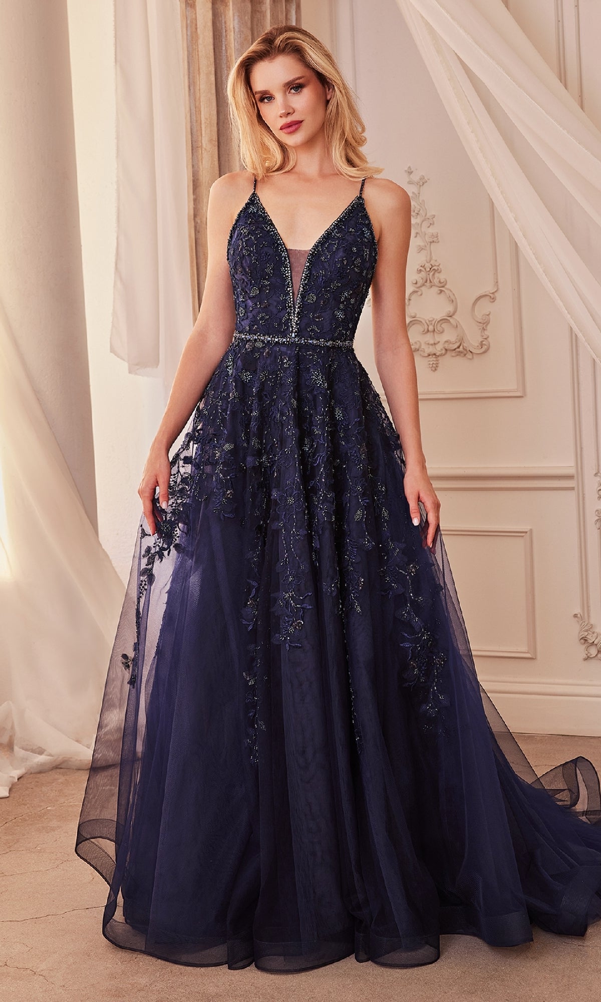 A woman stands in an elegant room wearing the Long Formal Dress A1251 by Andrea and Leo, a navy blue A-line ball gown with a deep V neckline and intricate floral embroidery that enhances her graceful look.