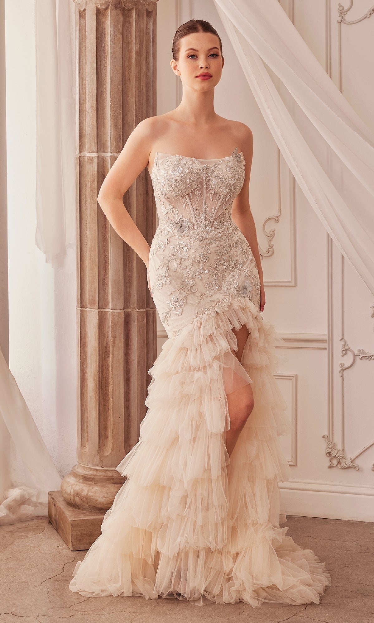 A woman stands indoors beside a large column wearing the Strapless Fitted Ruffle Prom Dress A1255, featuring intricate beading and a ruffled tulle skirt with a high slit.