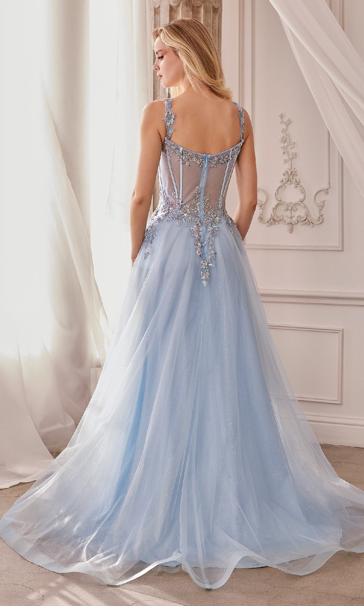 Woman in a Long Pastel A-Line Prom Dress (Andrea & Leo 1258), featuring a floor-length light blue embroidered gown with sheer back and appliqué details, standing indoors by draped curtains and ornate wall decor.