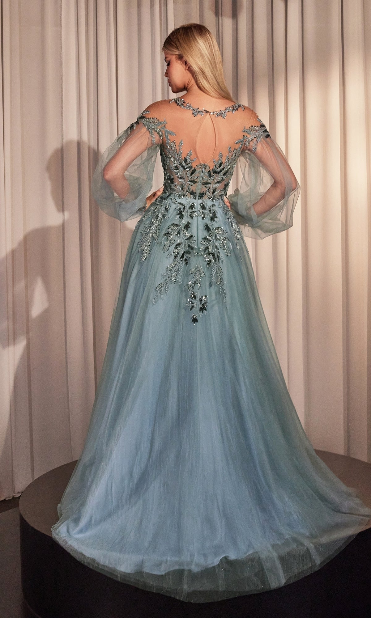 A woman in the Sheer Long Sleeve Long Formal Dress A1263, featuring a light blue hue and intricate floral embroidery, stands on a round platform facing away from the camera before pale curtains.