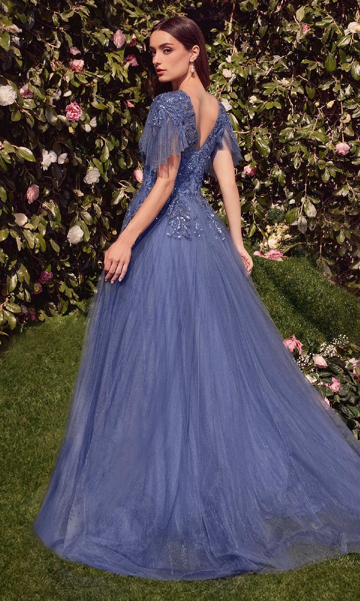 A woman stands outdoors before a leafy wall with pink flowers, wearing the Andrea & Leo A1351 short-sleeve prom ball gown—a blue tulle evening dress with lace details and an elegant floor-length a-line silhouette.