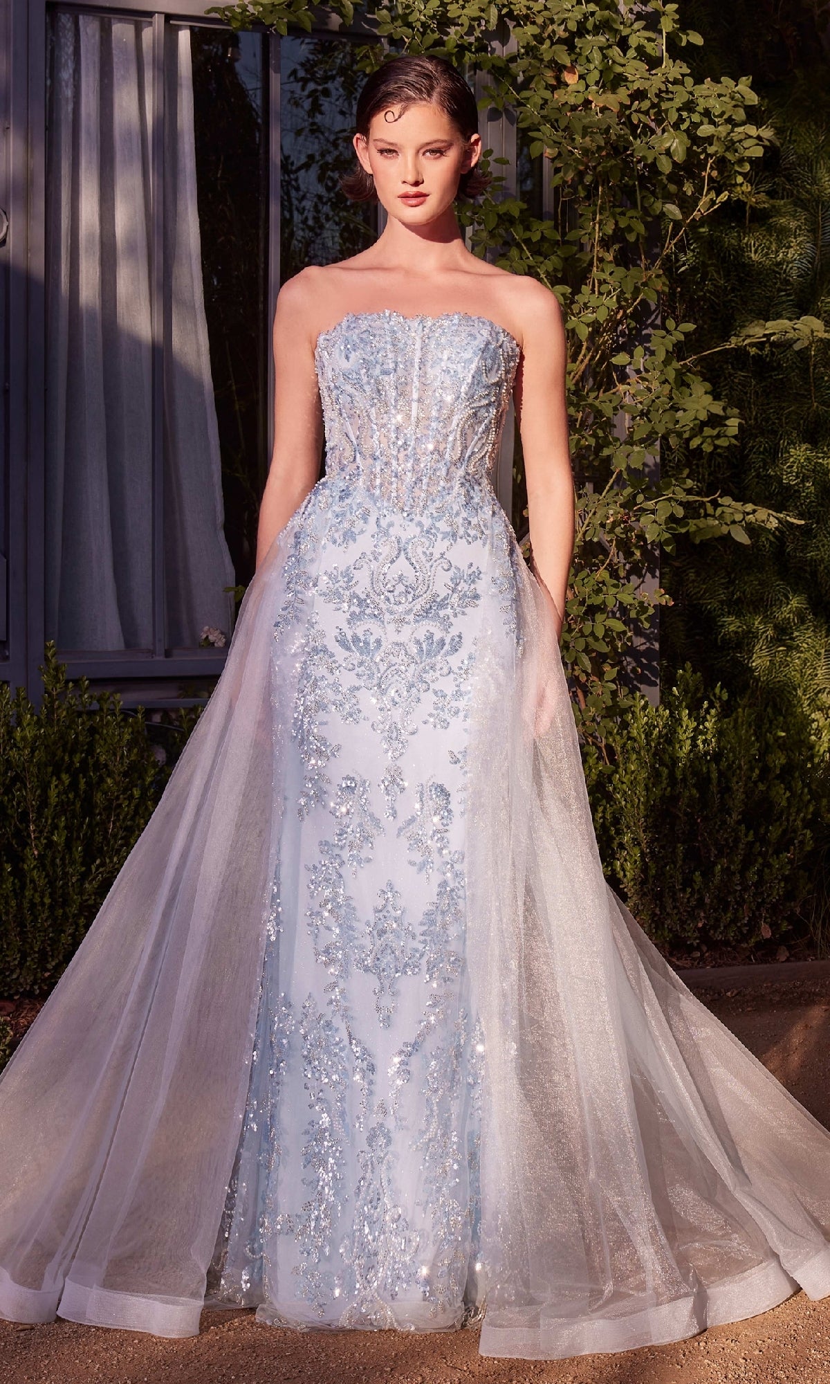 A woman wears the Andrea & Leo A1365 Strapless Sequin Prom Gown, featuring light blue sequins and a sheer tulle overlay, as she stands outside in front of greenery and a window.