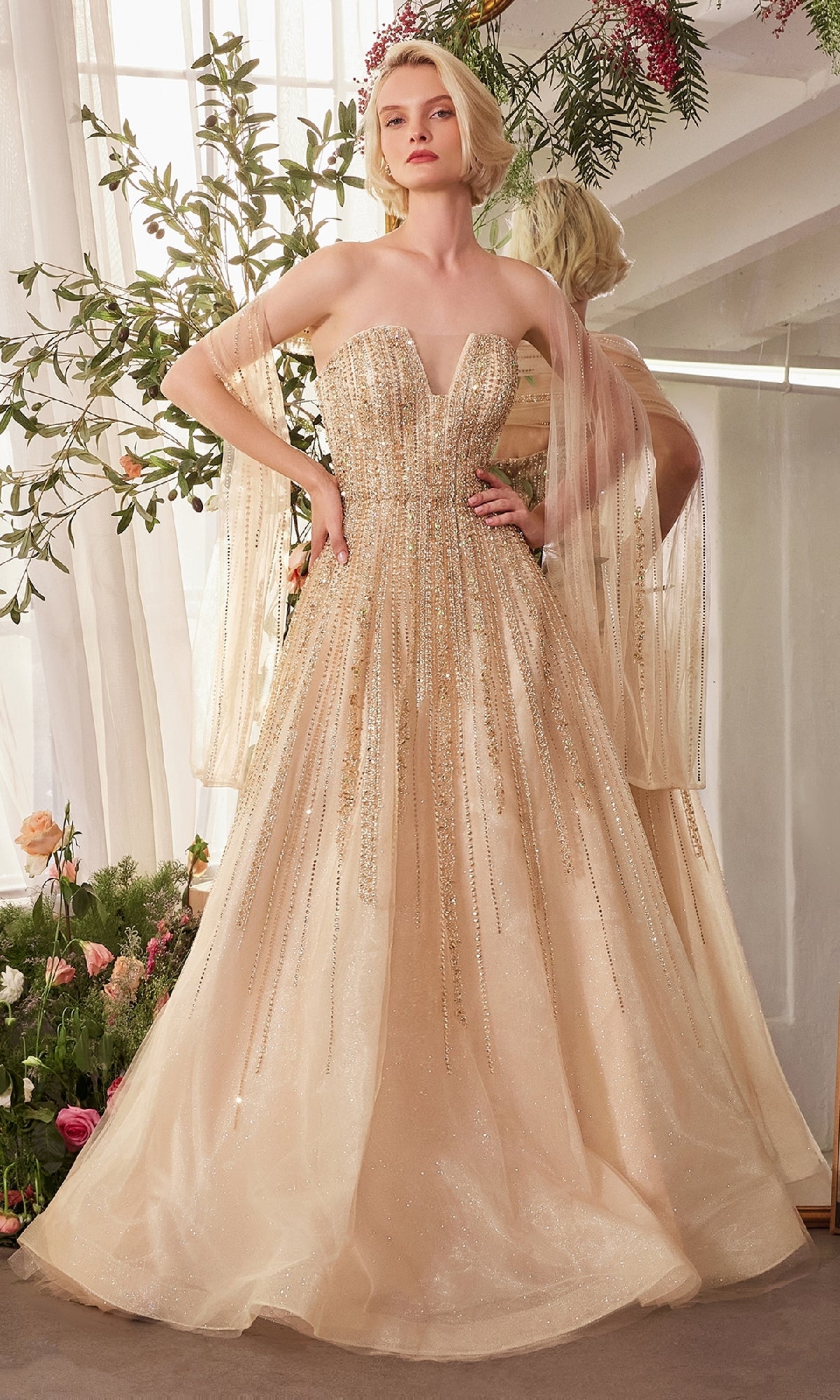 A woman stands indoors in the Andrea & Leo A1369, a strapless beaded long prom dress in sparkling champagne with sheer cape sleeves, surrounded by lush floral arrangements and greenery.