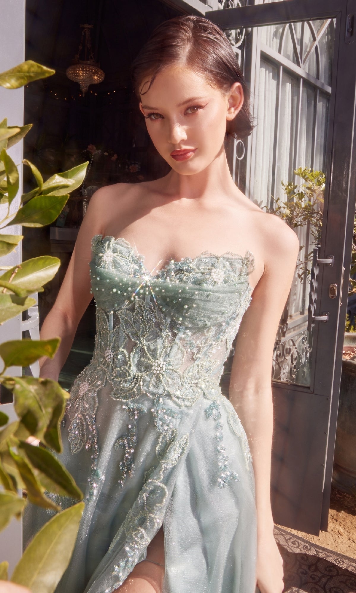 A woman in the Andrea & Leo A1378 Strapless Long Prom Dress with a corset-style waist stands by an open door, sunlight illuminating her and the lush greenery outside.
