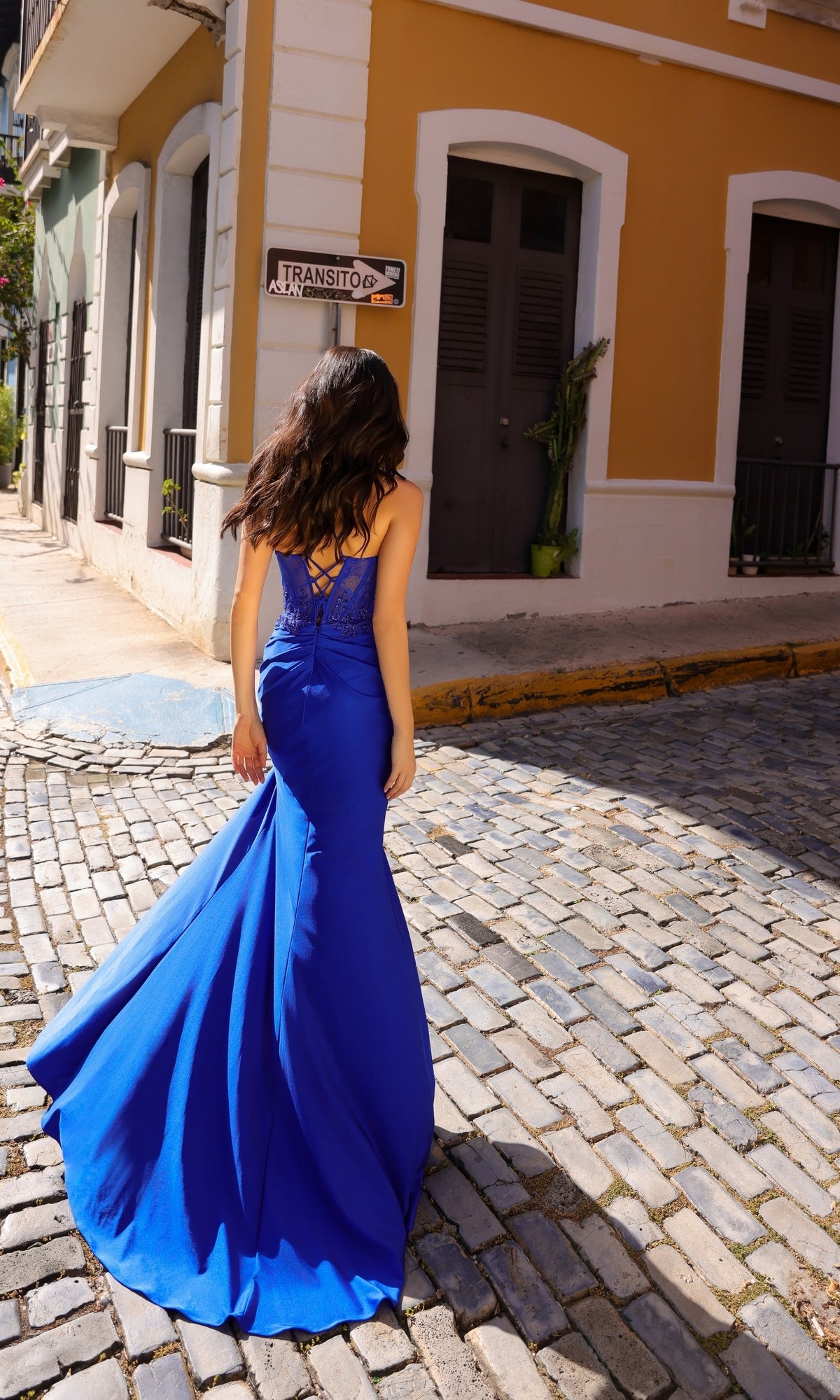 A woman wearing the Sheer-Beaded-Corset Long Prom Dress A1382 in bright blue walks on a cobblestone street by yellow and white buildings, facing away from the camera.