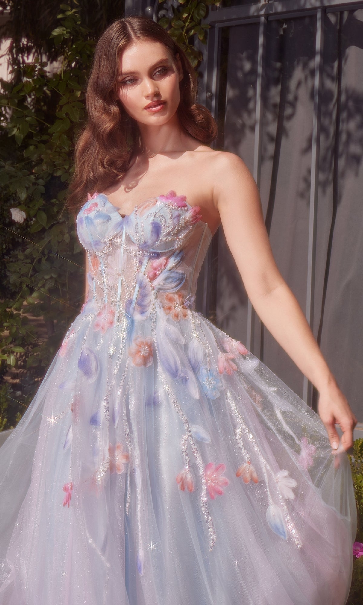 Outdoors, a woman wears the Long Strapless Prom Dress: Andrea & Leo A1383, featuring pastel floral appliqués and sheer tulle. Sunlight highlights her wavy hair and the gown’s delicate details.