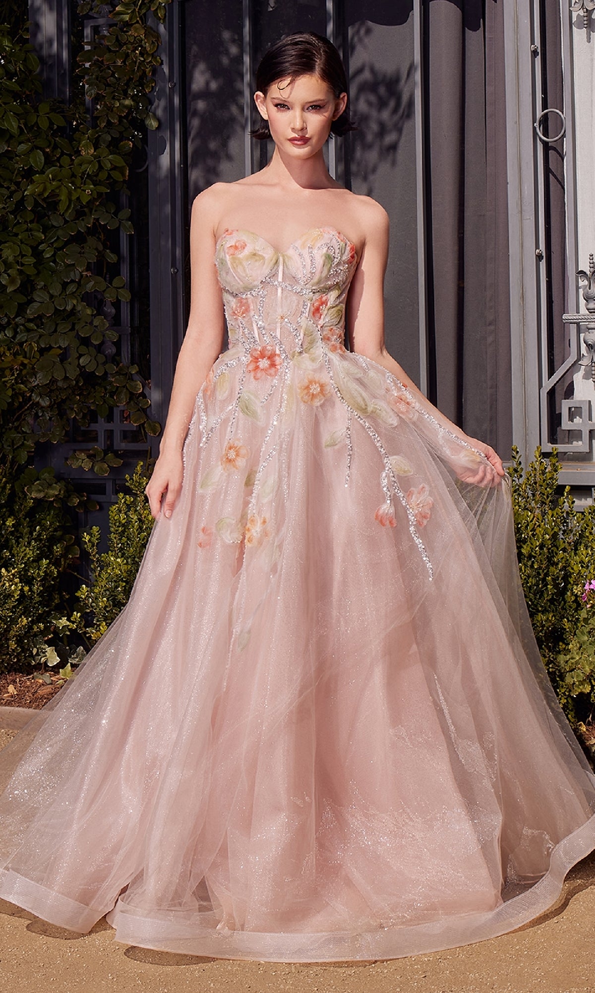 A woman stands outdoors in the Andrea & Leo A1383 long strapless prom dress, a light pink tulle gown with floral embroidery and a fitted bodice. Shrubs and architectural elements appear in the background.