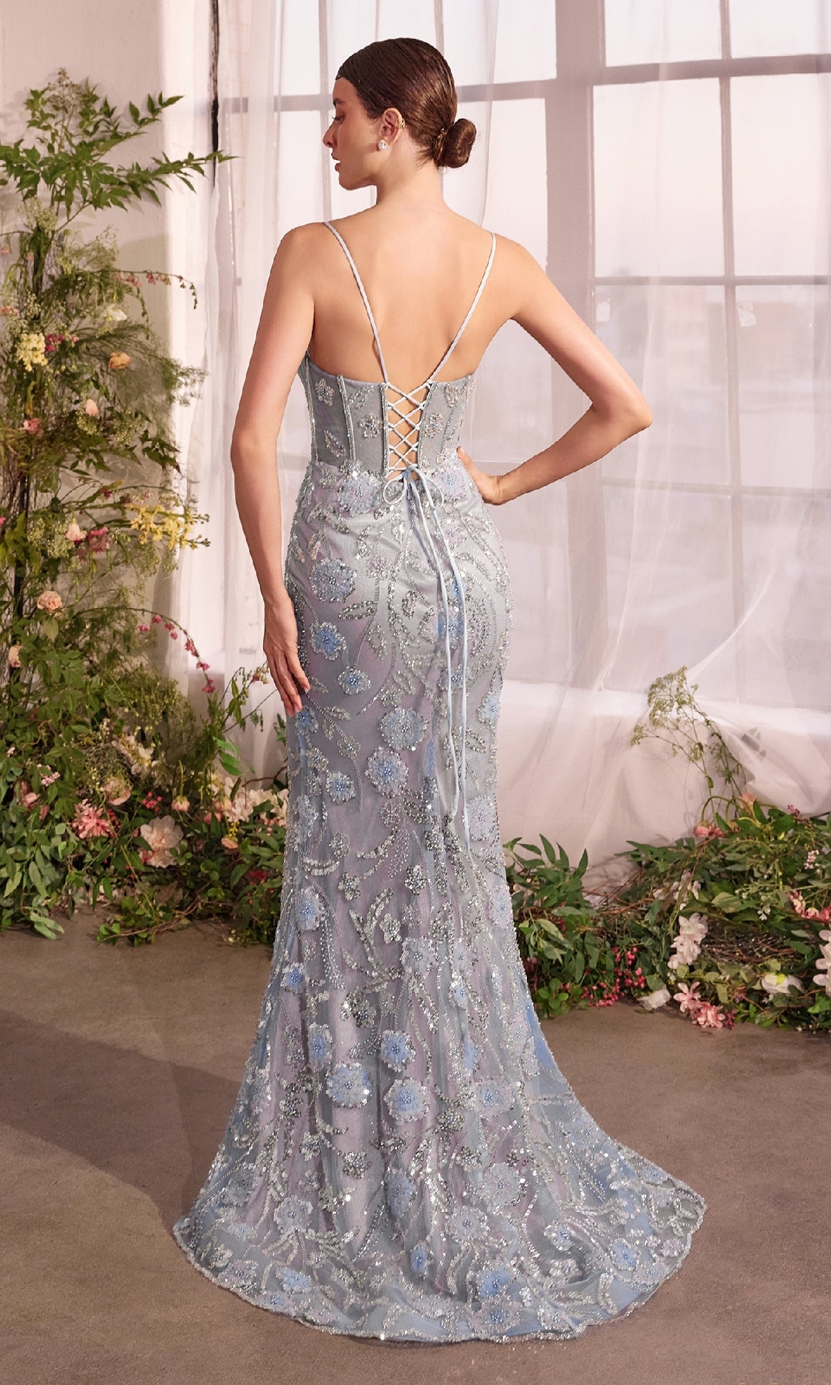 A woman stands indoors near large windows and greenery, wearing the Blue Floral Long Prom Dress: Andrea & Leo A1393—a light blue, floor-length mermaid gown featuring floral embroidery and a lace-up back that captures timeless elegance.