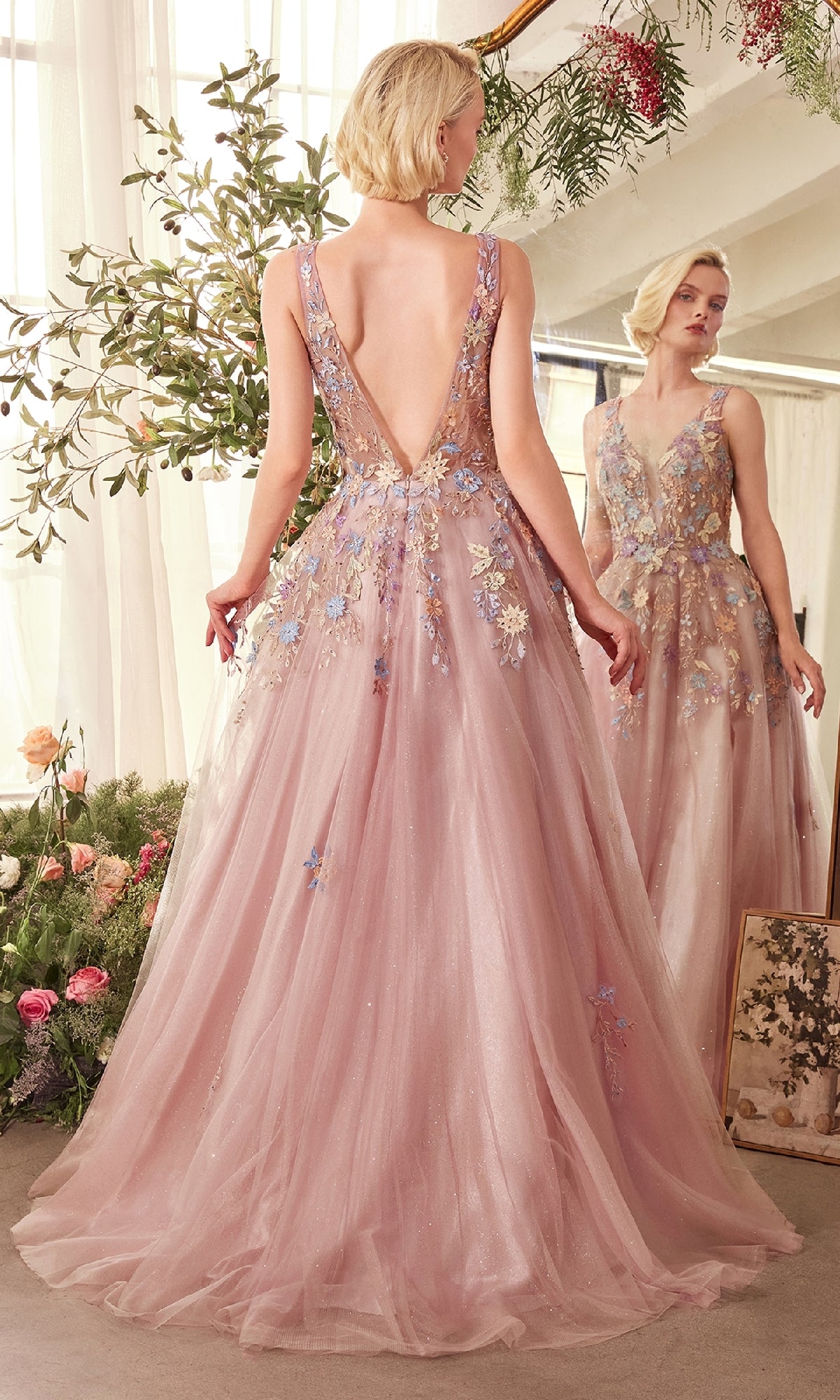 A woman wears the CLA1394 Mauve Pink Long Prom Ball Gown with floral-lace appliques and a deep V-back, standing before a mirror surrounded by flowers and greenery.
